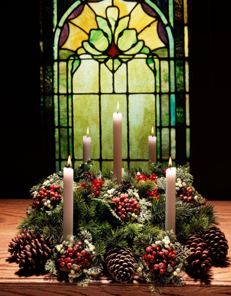 Corona de Adviento hecha con velas blancas, pino y bayas rojas frente a una ventana de cristal de opalino. | Foto: Getty Images