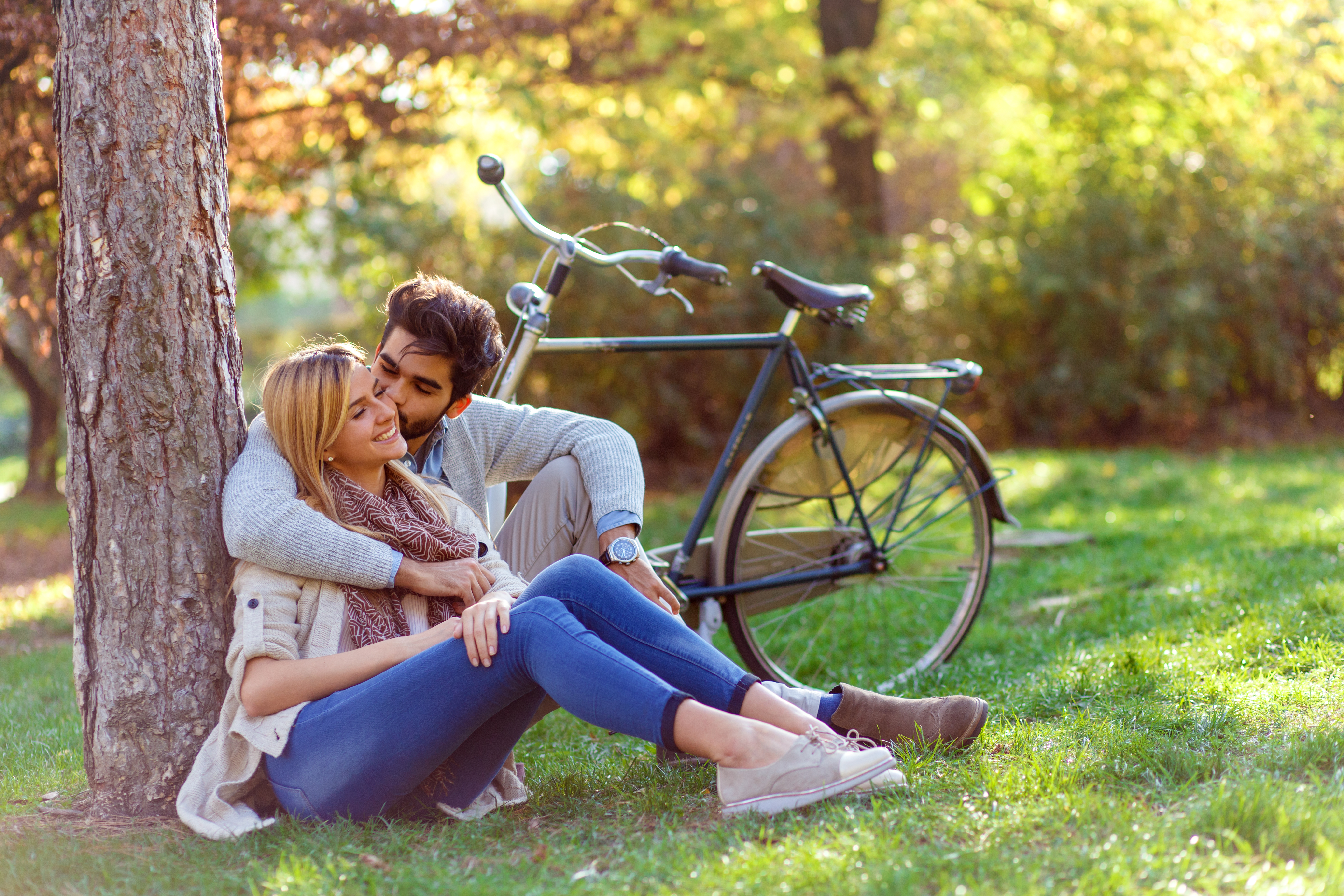 Una joven pareja de enamorados fotografiados sentados en un parque mientras se apoyan en un árbol y se abrazan. | Foto: Shutterstock