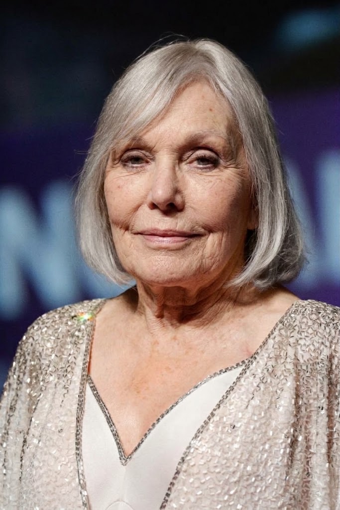 Kim Novak mira a la cámara con una expresión serena; su corte de pelo rubio plateado combina a la perfección con la brillante chaqueta plateada y el vestido blanco, todo ello sobre un fondo azul-morado ligeramente difuminado. | Fuente: Gemini