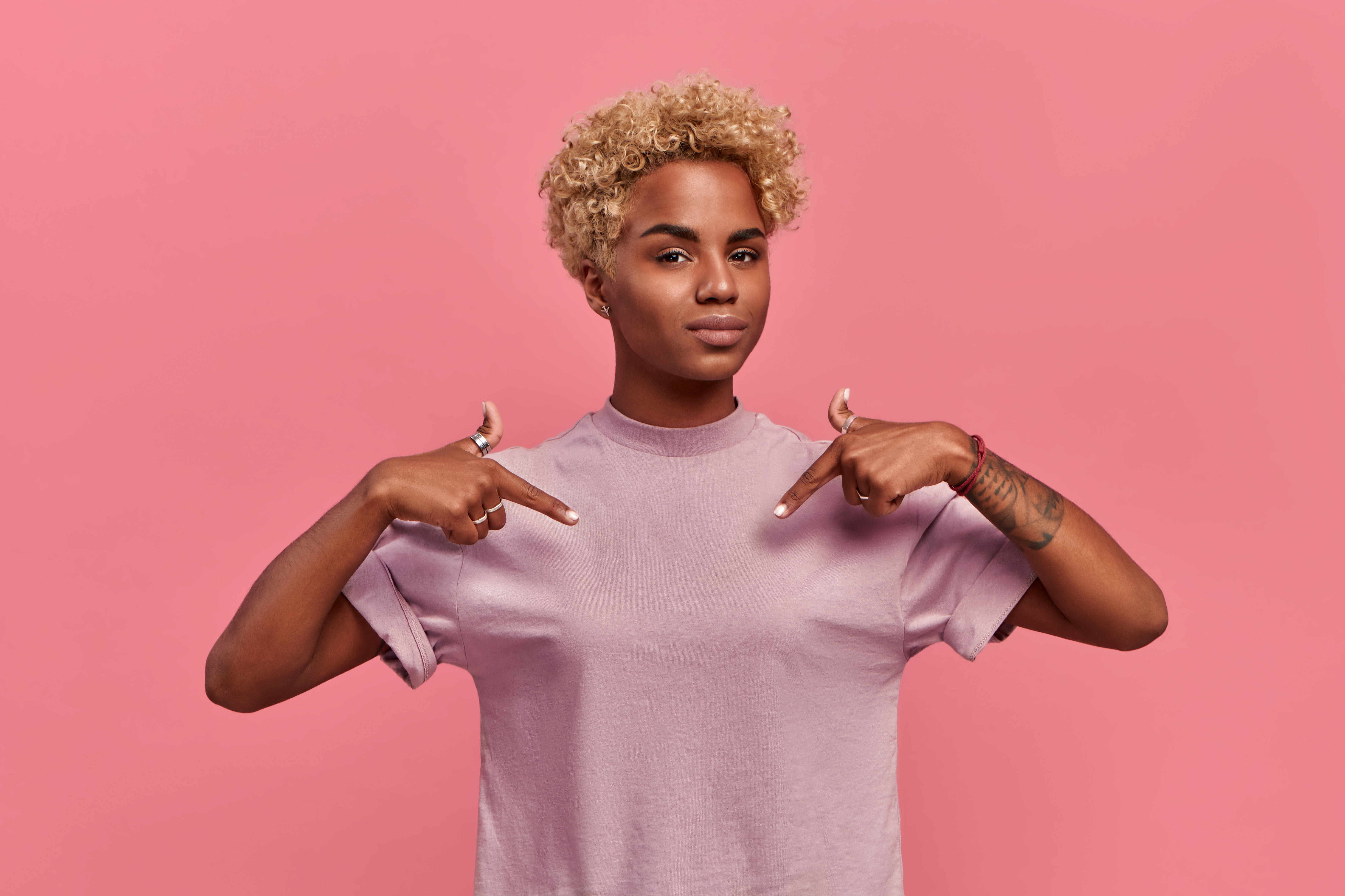Mujer afroamericana orgullosa y segura de sí misma, con peinado rubio y satisfecha de sus propios logros, se señala con las dos manos a sí misma o a una camiseta para imprimir, aislada sobre fondo rosa | Foto: Getty Images