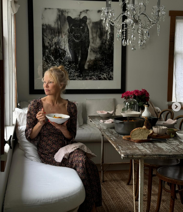 Con un vestido midi de flores y un moño, Pamela Anderson se relaja en un banco blanco acolchado en su rústico rincón de comedor de Arcady, sosteniendo un plato de sopa caliente mientras mira por la ventana. Una lujosa lámpara de araña de cristal cuelga sobre una mesa de madera envejecida, en la que hay pan recién hecho, rosas y ollas humeantes, todo ello bajo un espectacular cuadro en blanco y negro con motivos de la naturaleza. Compartió esta tranquila escena de la hora de la comida con el mensaje: "Descubre la belleza que hay en ti y luego compártela. 🤍". | Fuente: Instagram/pamelaanderson