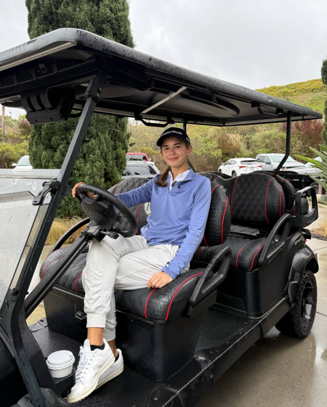 Kai sentado al volante de un carrito de golf, vestido con un top azul y pantalones blancos, sonriendo con una taza de café cerca | Fuente: Facebook/Kai-Trump