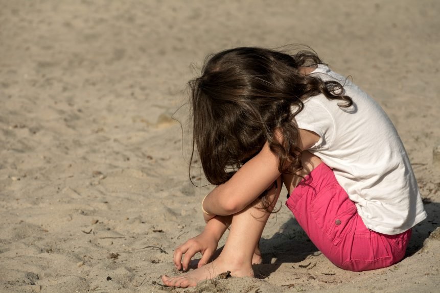 Niña sentada en el suelo con su rostro sobre las rodillas, jugando con la arena. | Imagen: Libreshot