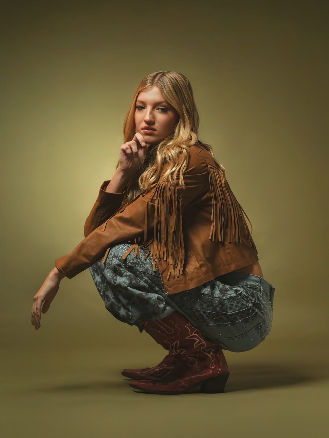 Allegra Cubero posa en cuclillas con chaqueta marrón con flecos, jeans estampados y botas, mirando a cámara