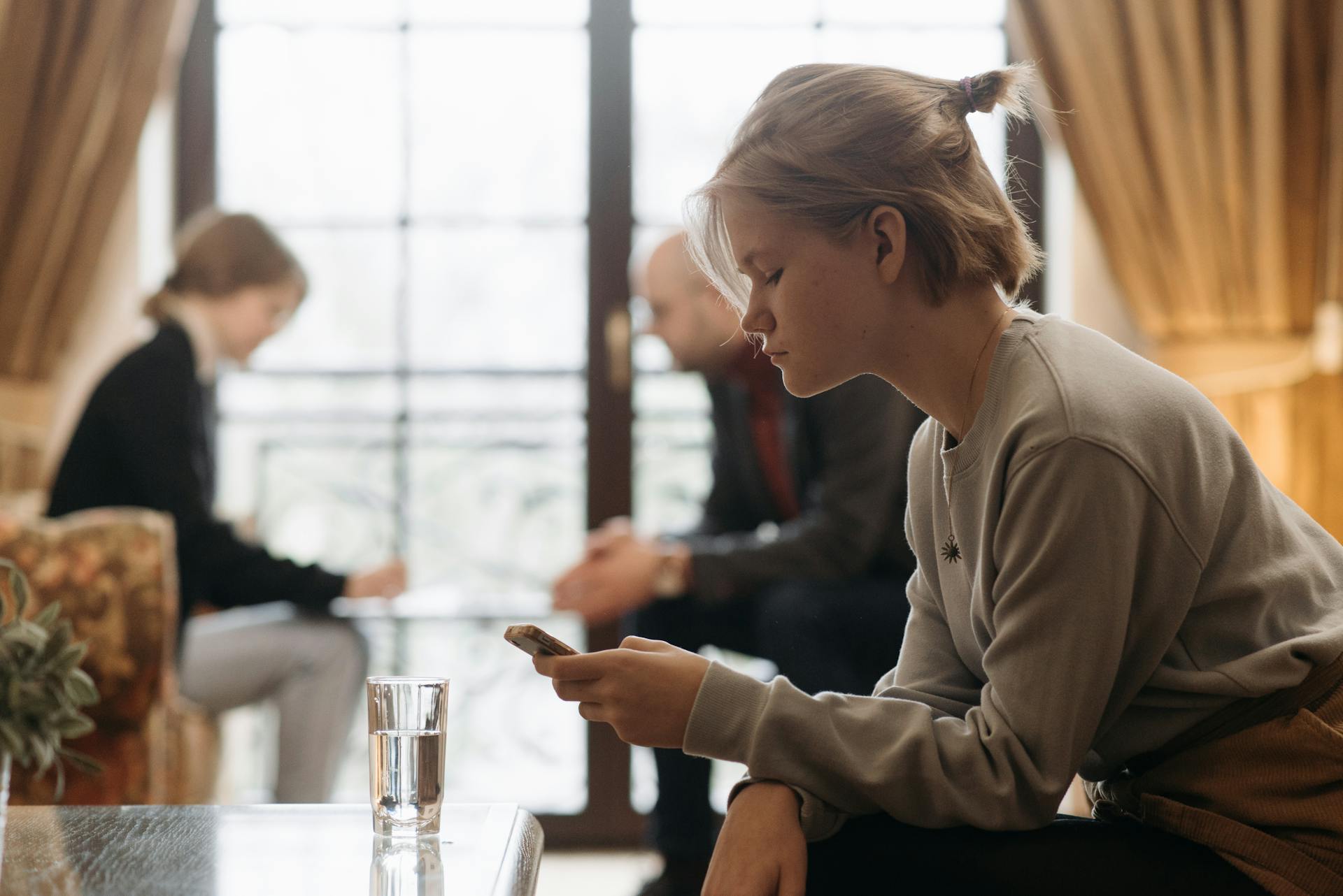 Una chica seria mirando su teléfono mientras otras personas aparecen en segundo plano | Fuente: Pexels