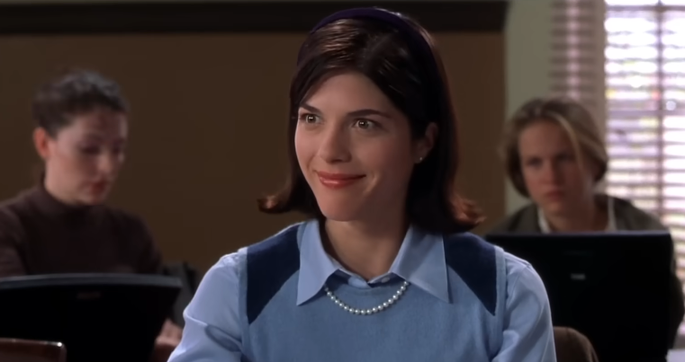 Selma Blair sonríe con aire de suficiencia desde su pupitre en el aula, interpretando a Vivian Kensington, en una escena de la película de 2001 "Legalmente rubia". Lleva una camisa preppy celeste con cuello bajo un chaleco de suéter azul claro con detalles azul marino en los hombros, y accesorios como un clásico collar de perlas, aretes pequeños y una diadema oscura que sujeta su bob castaño peinado hacia afuera. | Fuente: YouTube/Amazon MGM Studios