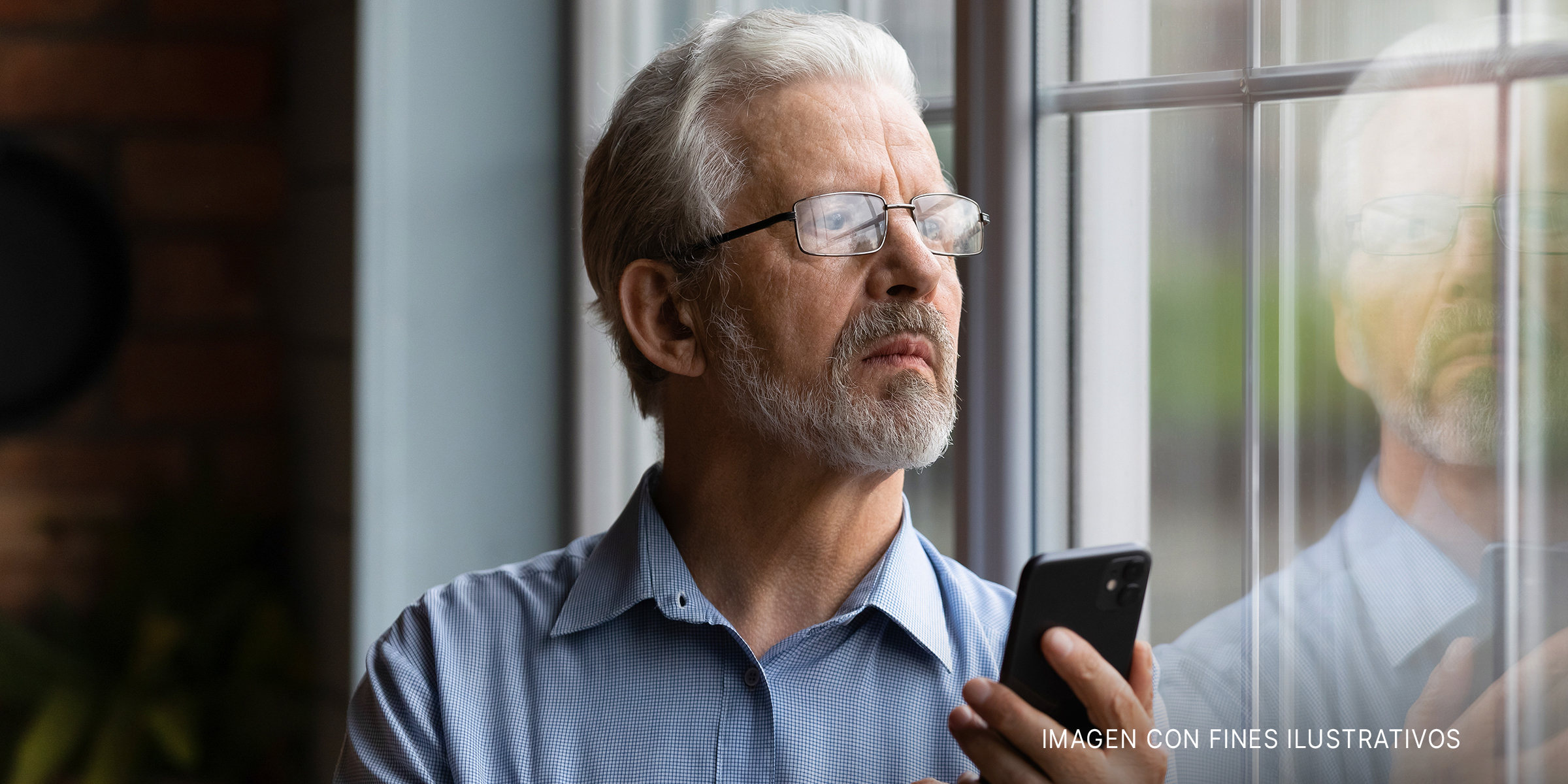 Un hombre mayor sujetando un teléfono de pie y mirando por la ventana | Foto: Shutterstock