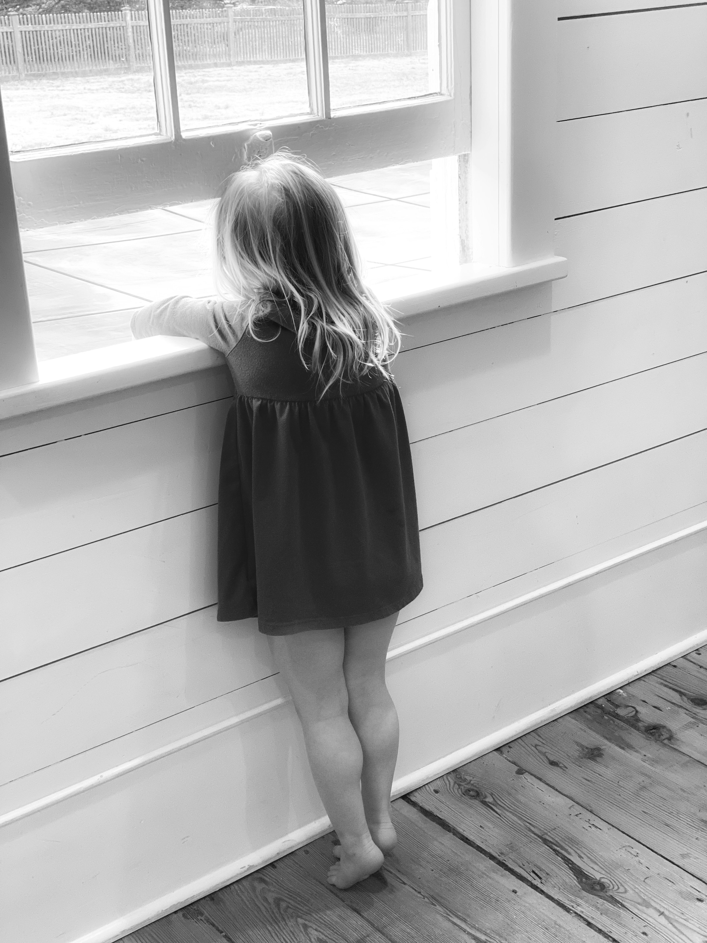 Una toma en escala de grises de una niña descalza mirando por la ventana | Fuente: Unsplash