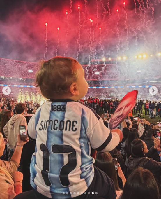 Desde una toma de espaldas sobre los hombros de su padre, Faustino contempló el impactante show de fuegos artificiales y luces que iluminó el cielo del estadio. El bebé lució la camiseta argentina personalizada con el apellido Simeone y el dorsal 17, sosteniendo la pancarta con el rostro de Messi mientras el humo tiñó de rojo el ambiente festivo. | Fuente: Instagram/evabargiela