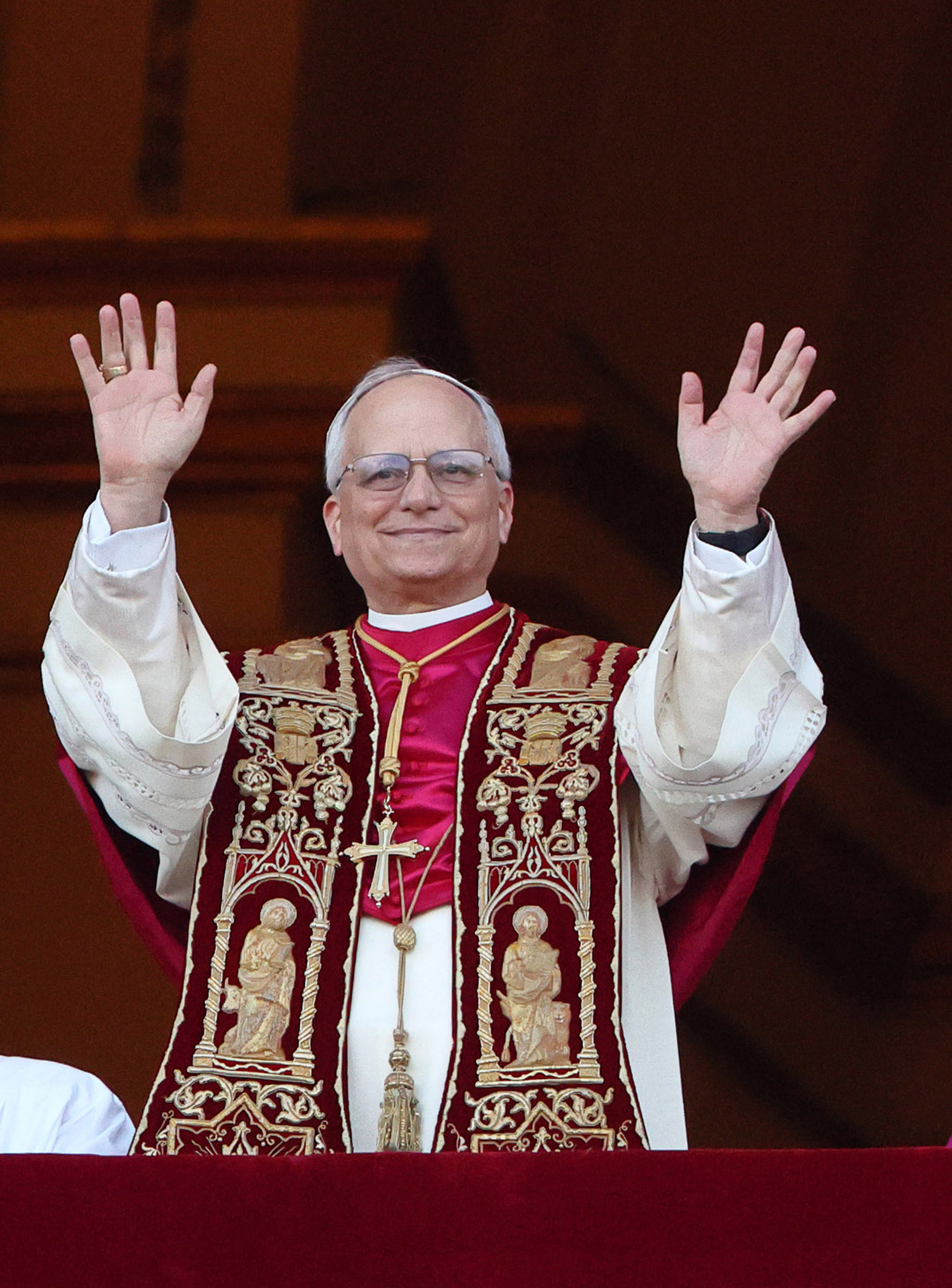 El papa León XIV sonríe cálidamente mientras levanta ambas manos en señal de saludo, su ornamentada estola carmesí detallada con santos dorados capta la luz