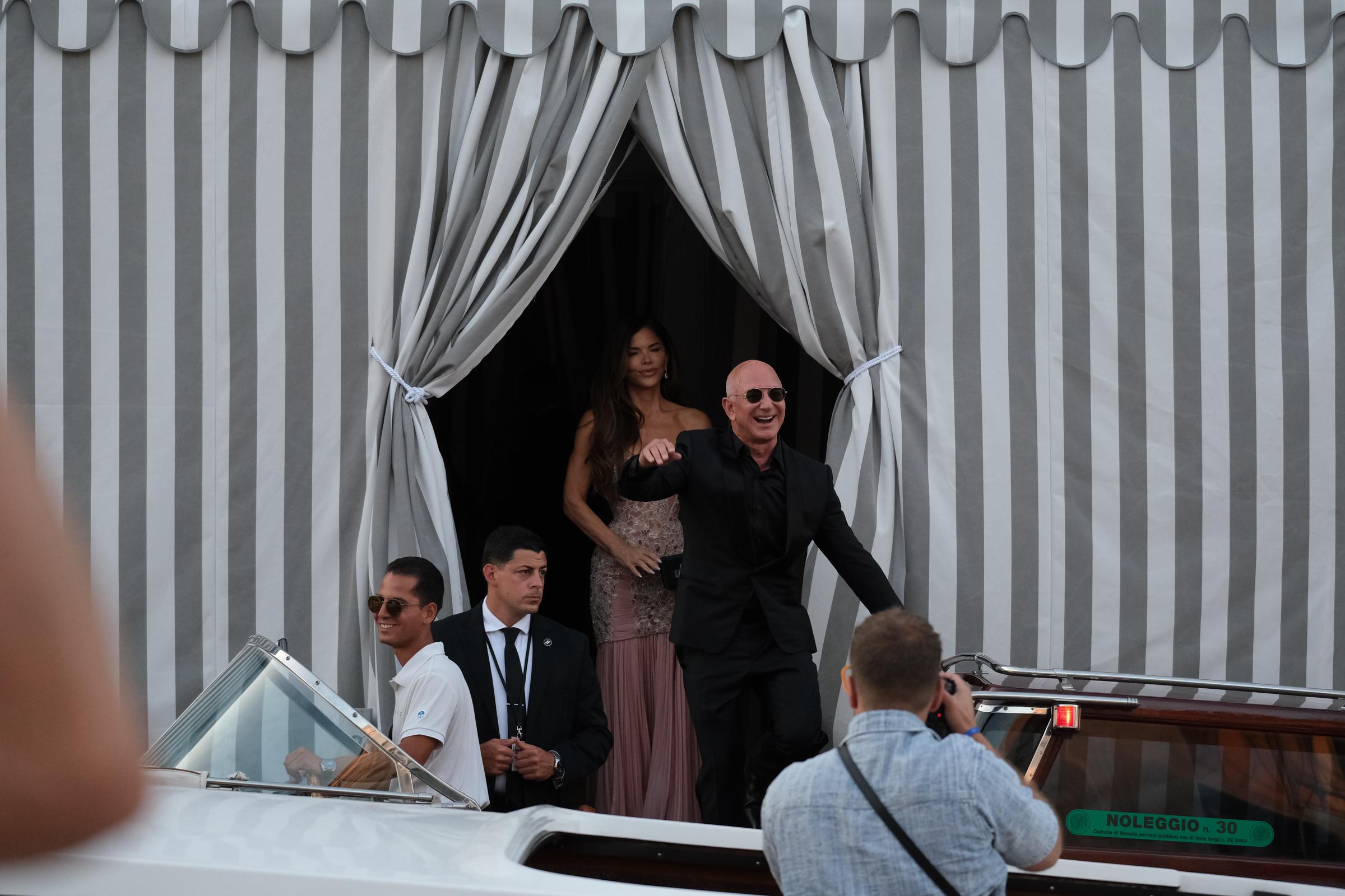 Jeff Bezos sonríe y saluda con entusiasmo mientras sube a un taxi acuático, mientras Lauren Sánchez le sigue con un vestido de cuentas en tonos rubor, enmarcado por cortinas de rayas y rodeado de personal y curiosos.