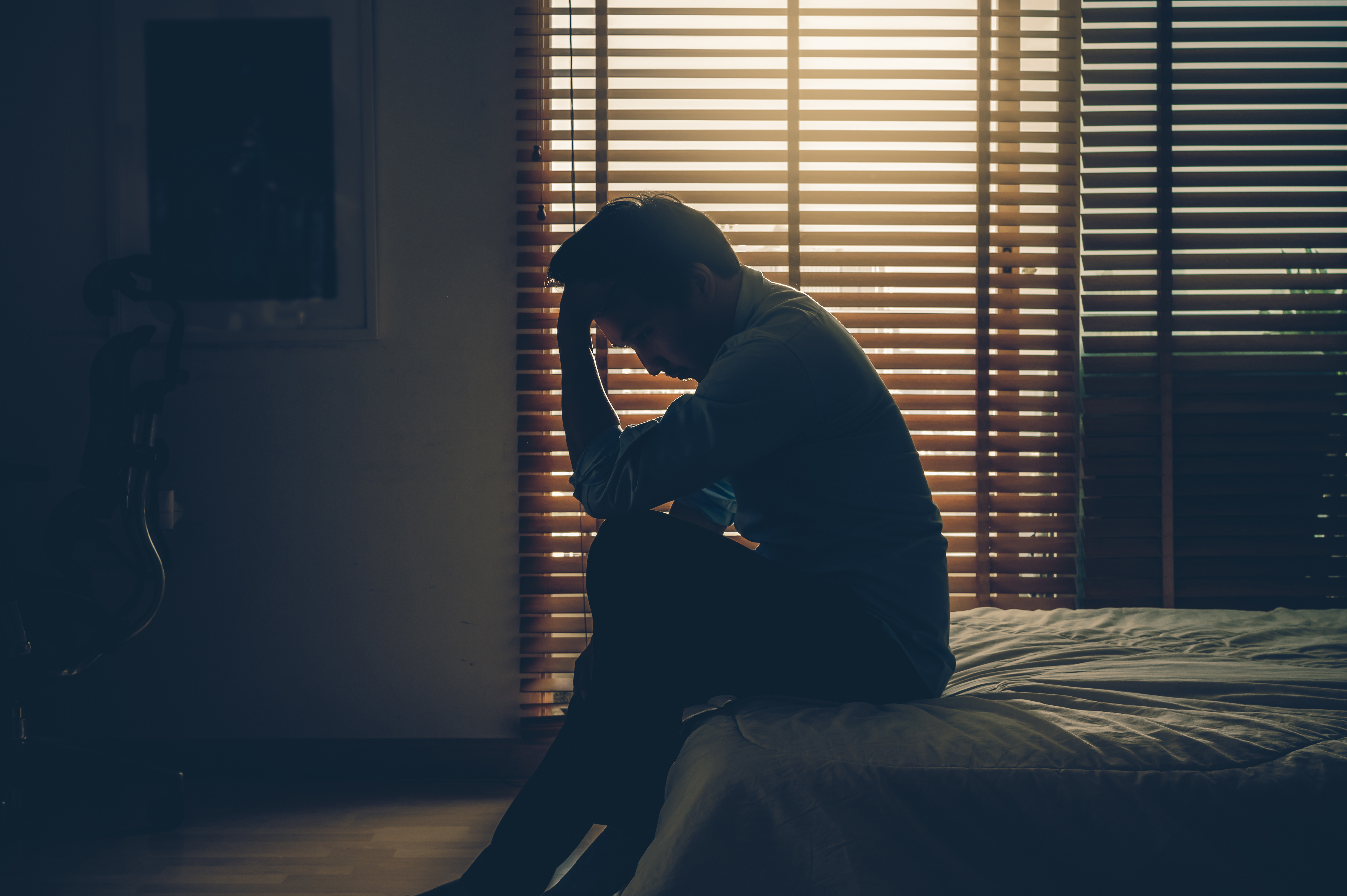 Hombre de negocios triste sentado con la cabeza entre las manos sobre la cama en un dormitorio oscuro con poca luz ambiental. | Fuente: Shutterstock