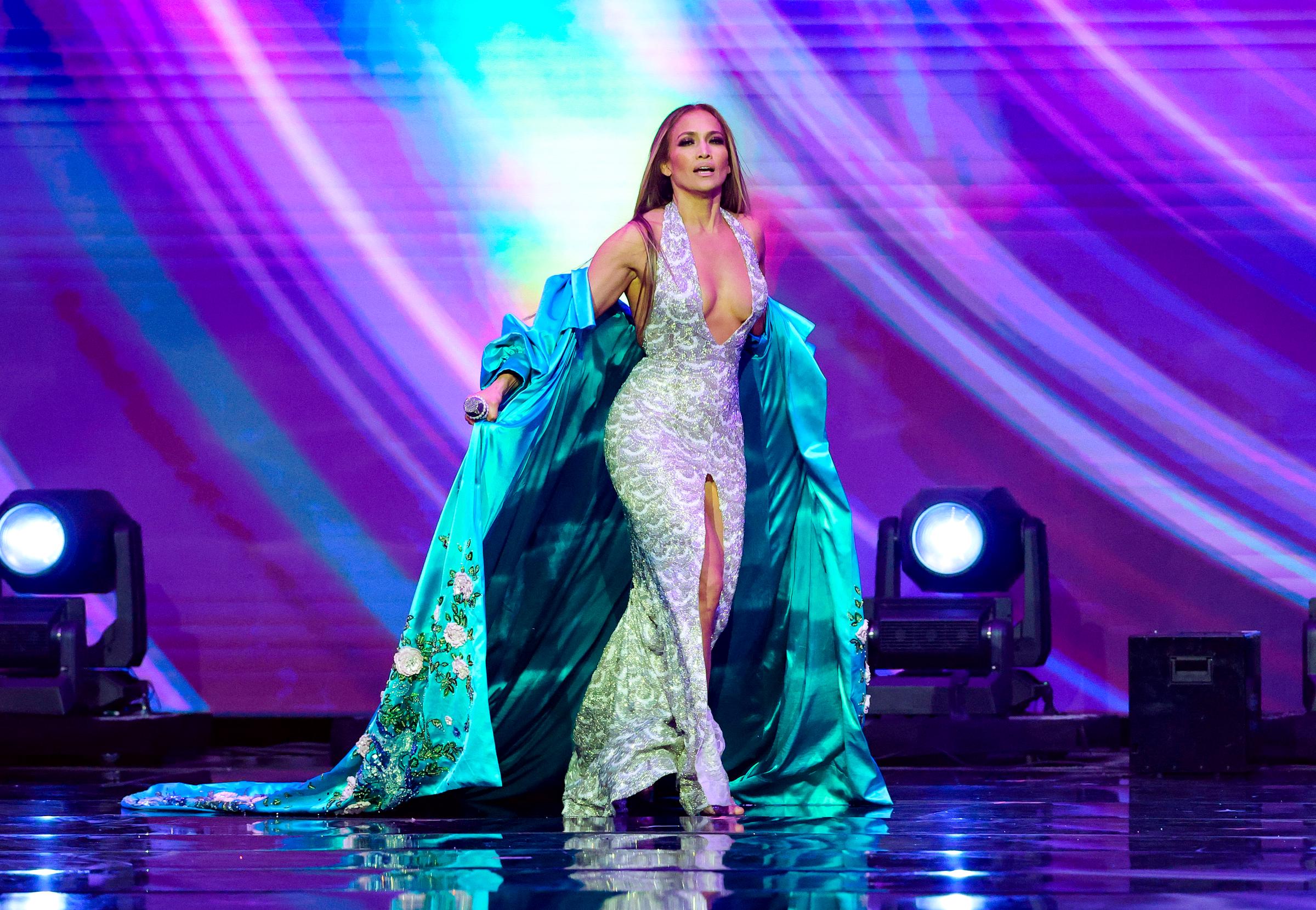 J.Lo cruza el escenario con confianza, abriéndose la bata para mostrar el brillo de su vestido plateado bajo las deslumbrantes luces.