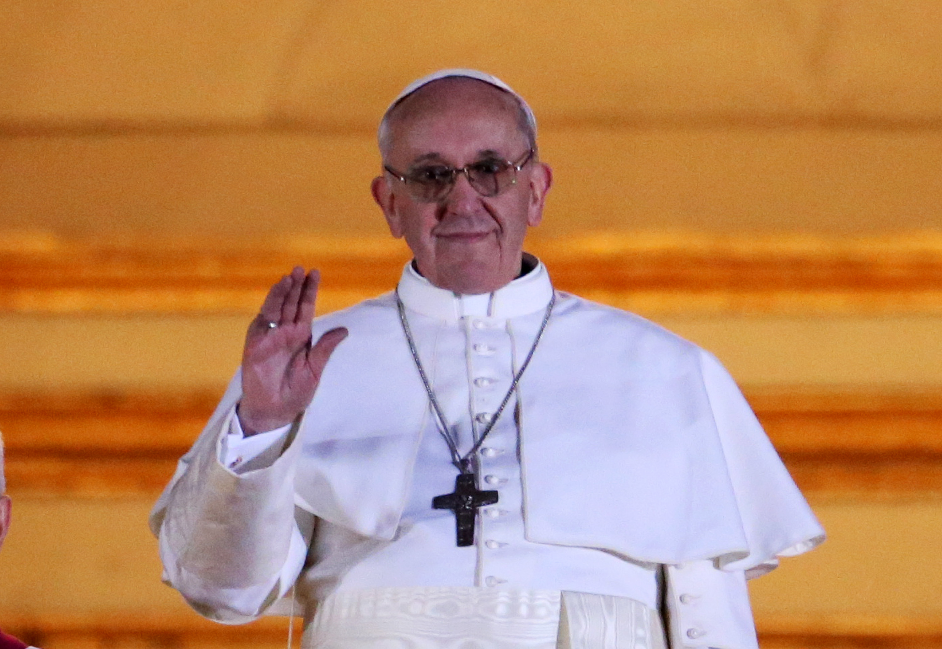El papa Francisco saluda a la multitud con un suave gesto de la mano, vestido con una sencilla túnica blanca y una cruz de plata, con una sonrisa tranquila enmarcada por una suave luz dorada.