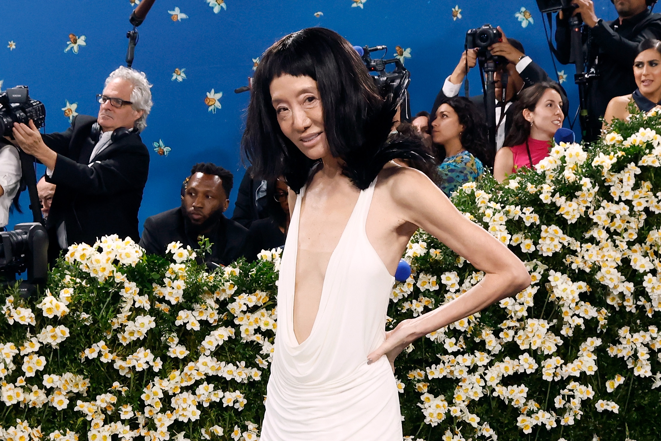Posando con confianza, Vera Wang se alza sobre un campo de flores blancas, resaltando el escote pronunciado y los hombros de plumas de su vestido hecho a medida.