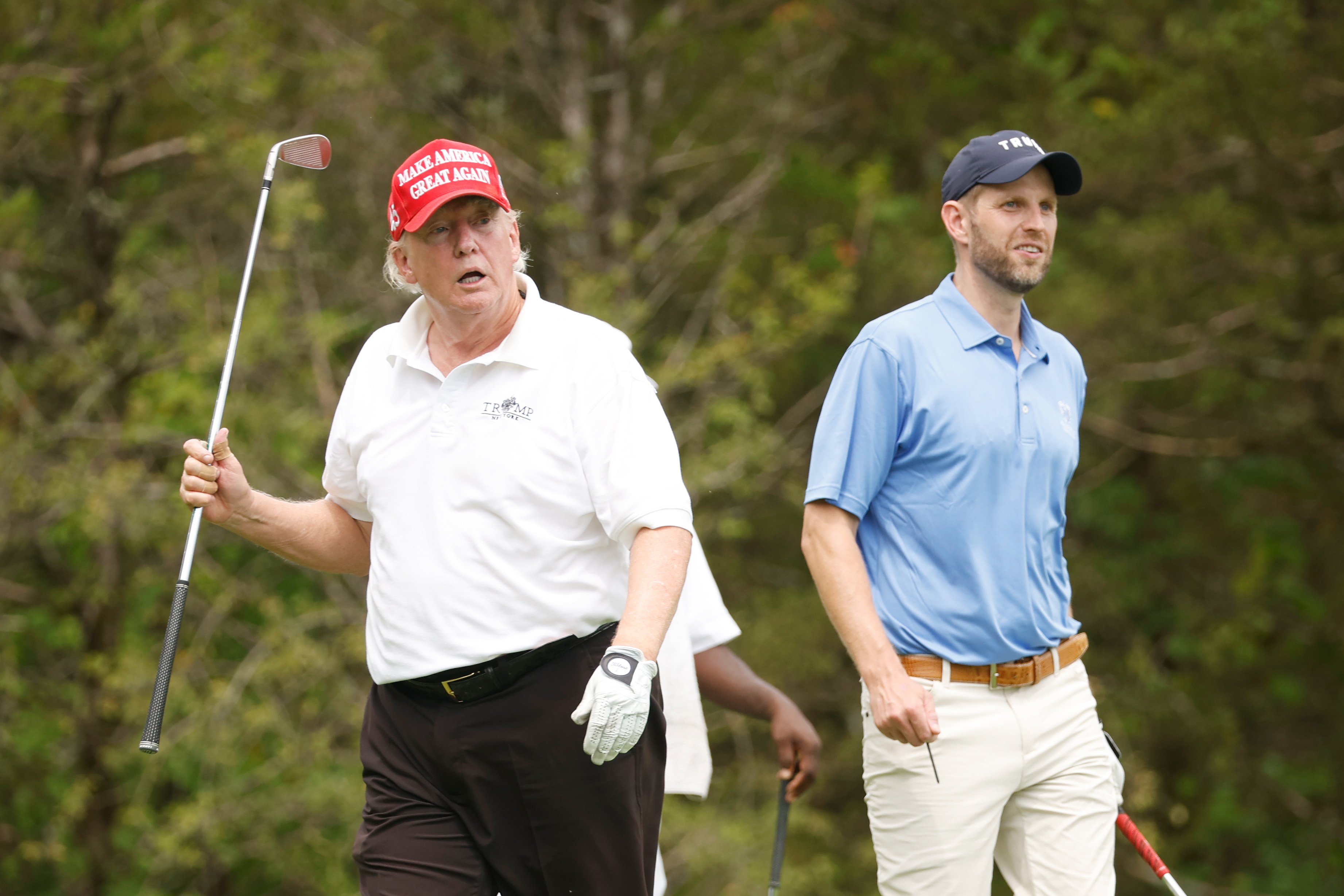 Donald Trump camina junto a su hijo, Eric Trump, durante el torneo, sosteniendo un palo en el aire. Su rostro muestra un enrojecimiento visible alrededor de las mejillas y la nariz, mientras que el resto de su piel parece más clara.