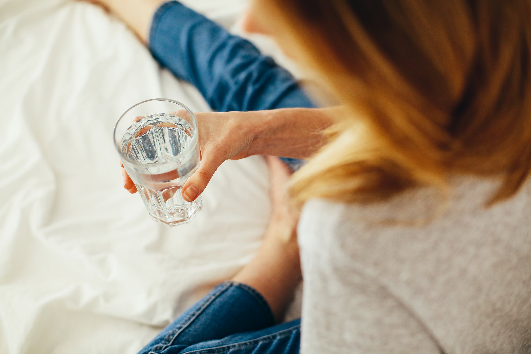 Mujer sosteniendo un vaso con agua mientras está sentada en una cama. | Foto: Pexels
