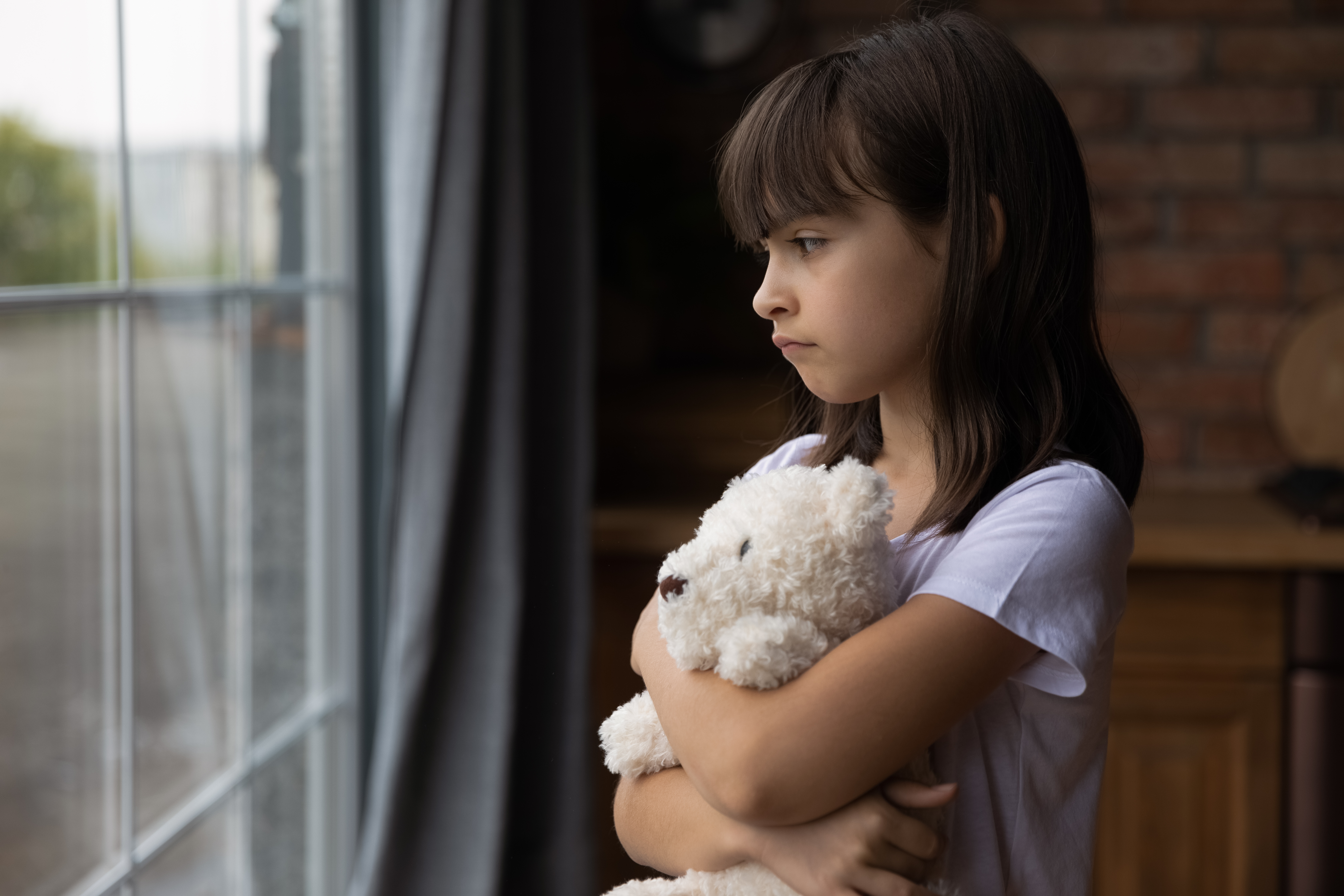 Una niña mirando por la ventana mientras abraza un juguete. | Foto: Shutterstock