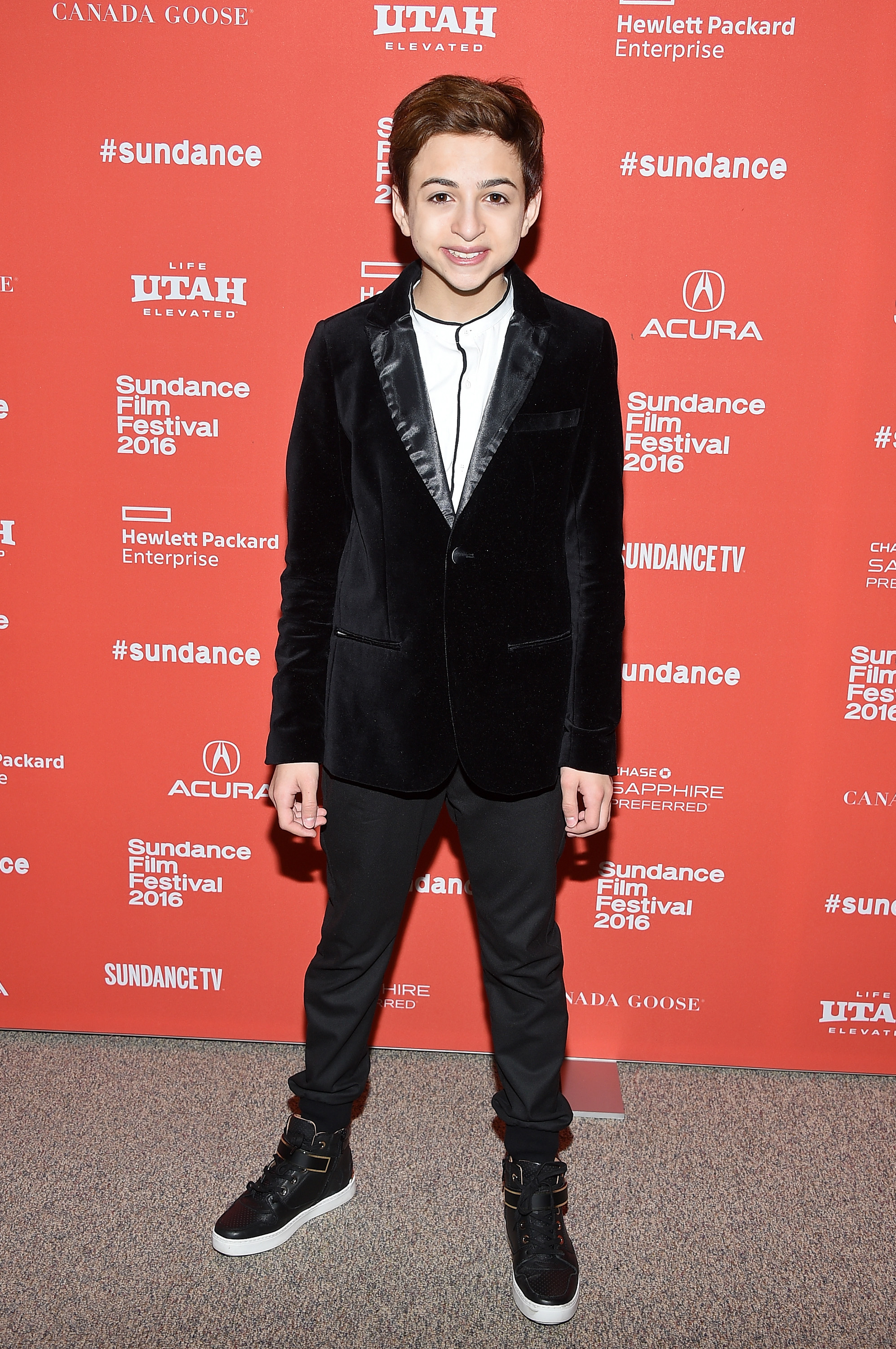 J.J. Totah sonríe a los fotógrafos en el estreno de "Other People" durante el Festival de Cine de Sundance en Park City, Utah, vestido con una chaqueta de terciopelo negro, una camisa blanca, pantalones negros y zapatillas de caña alta. | Fuente: Getty Images