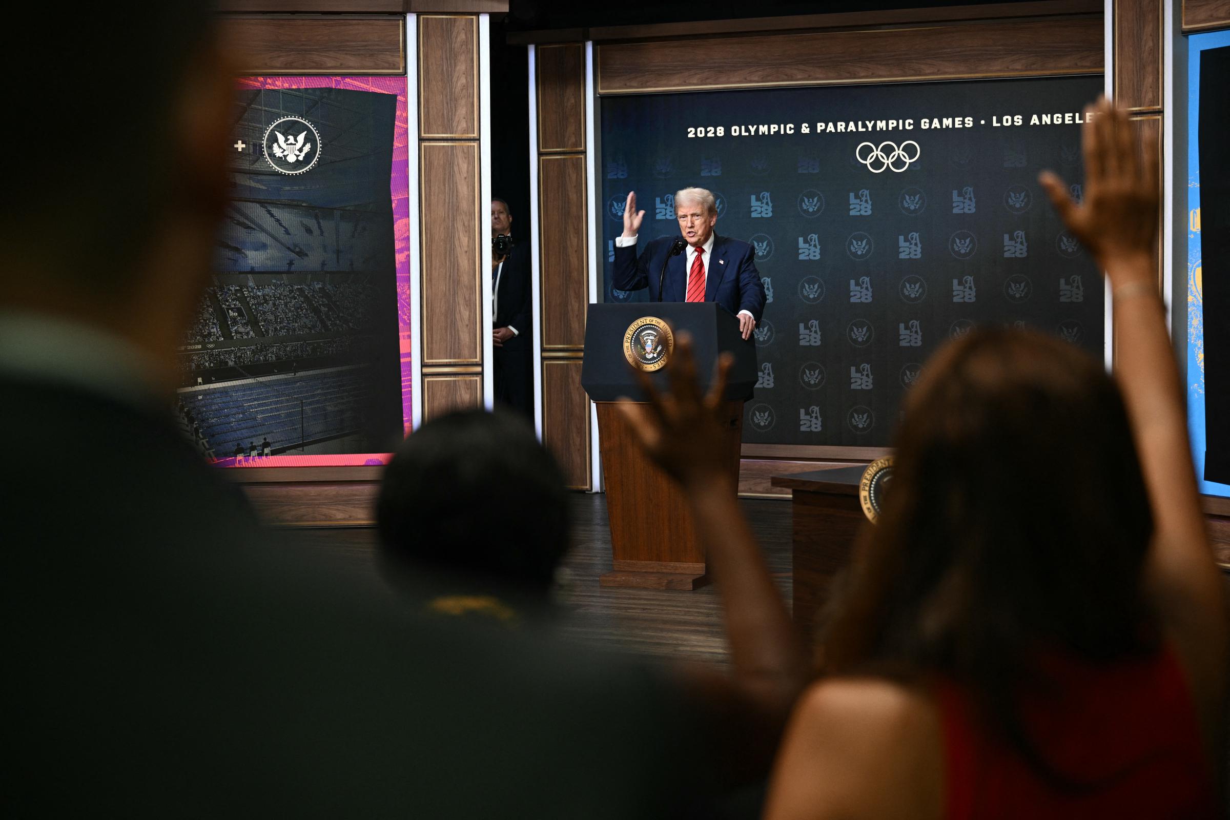 Tras su paseo por la azotea de la Casa Blanca, Donald Trump habla desde el podio del Auditorio South Court, respondiendo a las preguntas tras firmar una orden ejecutiva para formar un grupo de trabajo sobre los Juegos Olímpicos de 2028, mientras los miembros de la prensa levantan la mano.