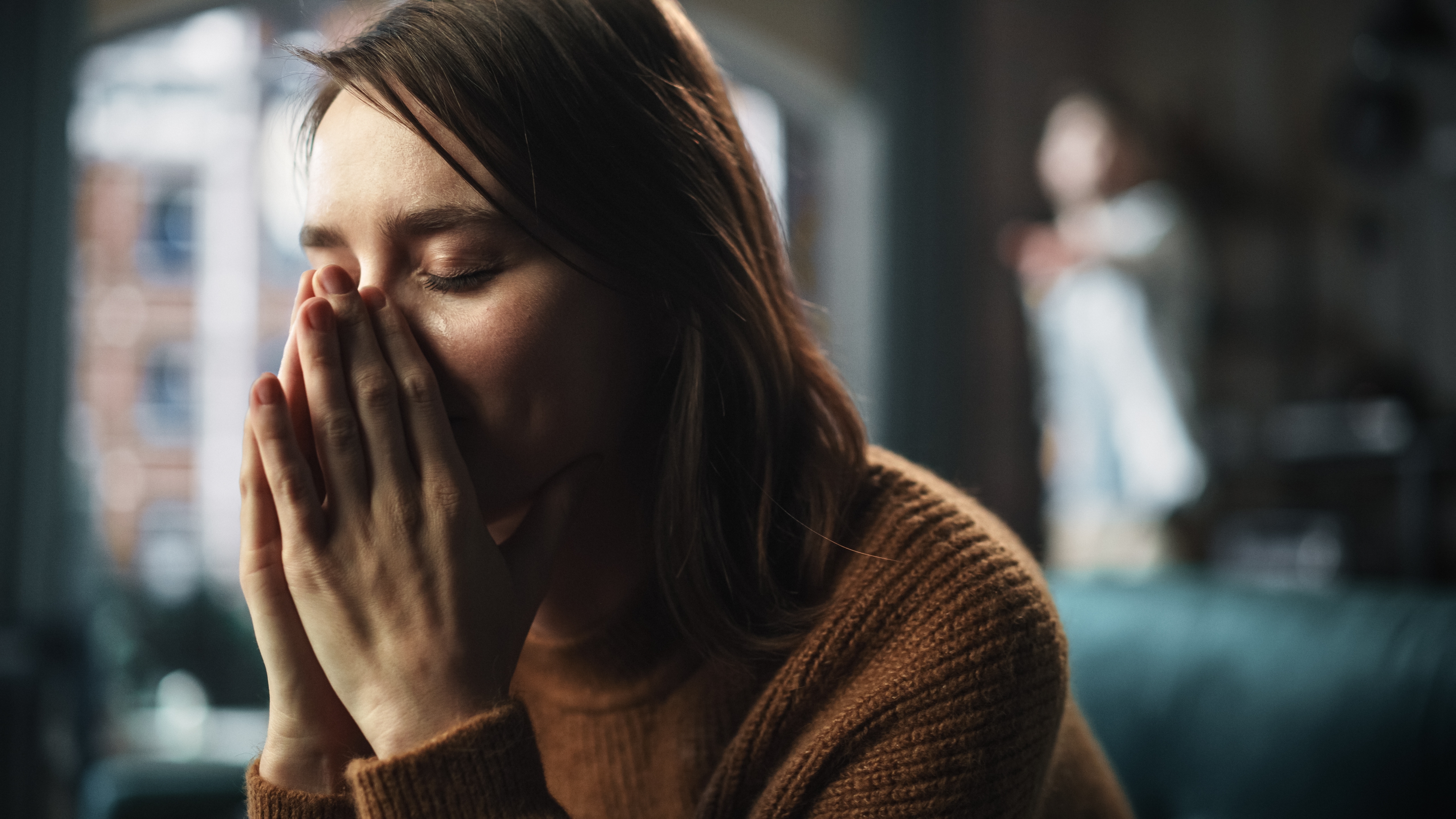 Una mujer disgustada se tapa la cara mientras está sentada en un sofá con alguien de pie al fondo. | Foto: Shutterstock