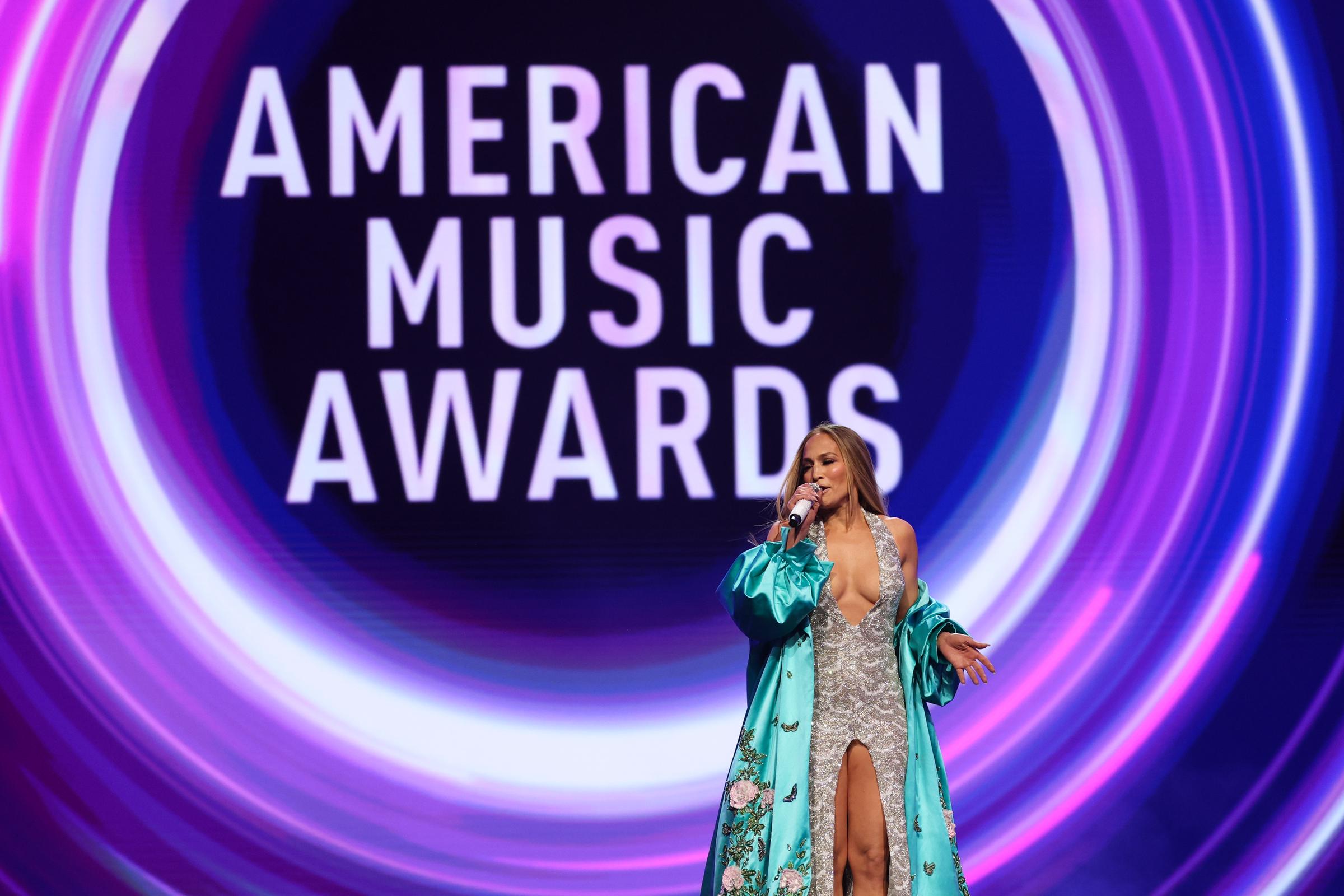 A mitad de la presentación, Jennifer Lopez levanta una mano mientras habla, el brillo de su vestido y el movimiento de su bata bordada destacan sobre los atrevidos y arremolinados gráficos del escenario de los AMAs.
