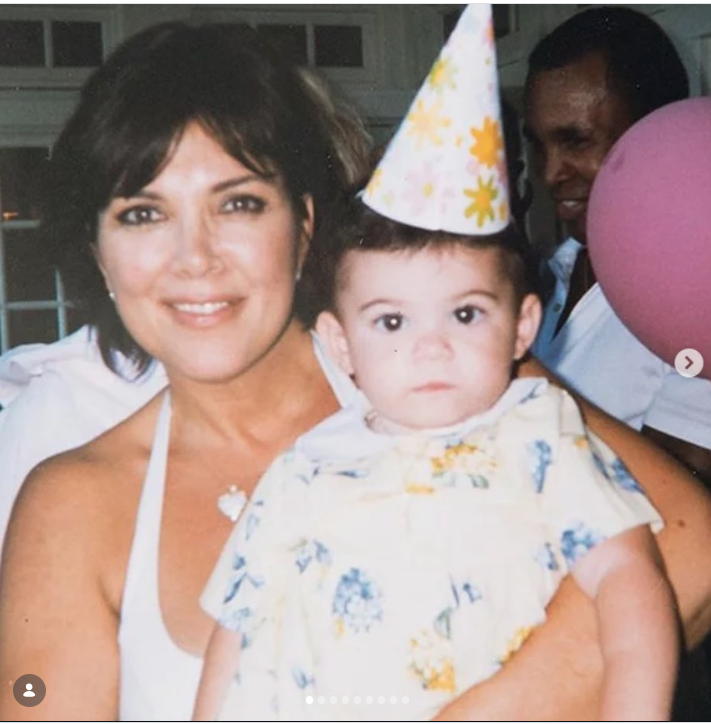 Una joven Kris Jenner sostiene en brazos a la pequeña Kylie Jenner durante una fiesta de cumpleaños; Kylie lleva un conjunto de flores y un gorrito de fiesta. Kris sonríe a la cámara mientras acuna a su hija menor en sus brazos. | Fuente: Instagram/krisjenner