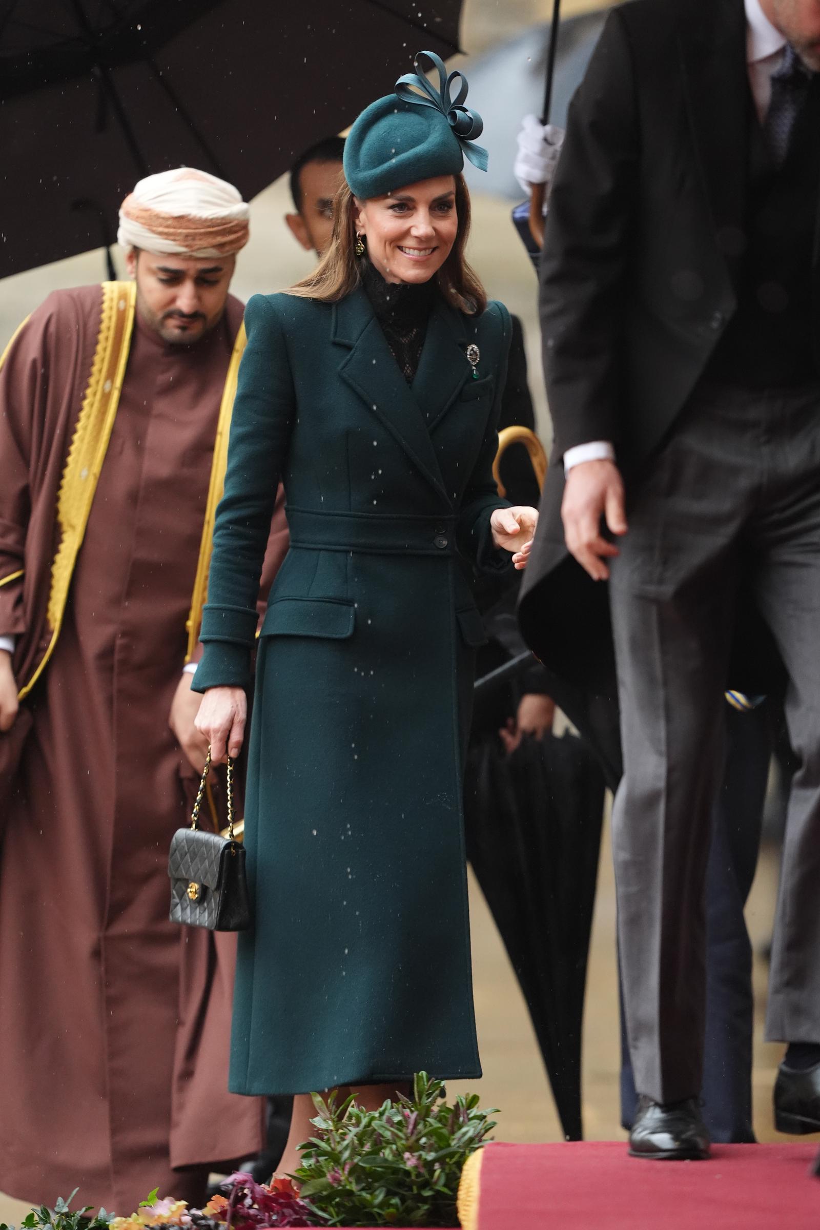 Catherine, princesa de Gales, sonríe mientras camina por el suelo húmedo del recinto ceremonial de Windsor el 20 de octubre de 2026. Viste un abrigo entallado verde bosque sobre una delicada prenda de encaje negro de cuello alto, con el histórico broche de las tres plumas de la reina Alexandra en la solapa. Lleva un pequeño bolso Chanel negro acolchado mientras camina cerca del príncipe William. | Fuente: Getty Images