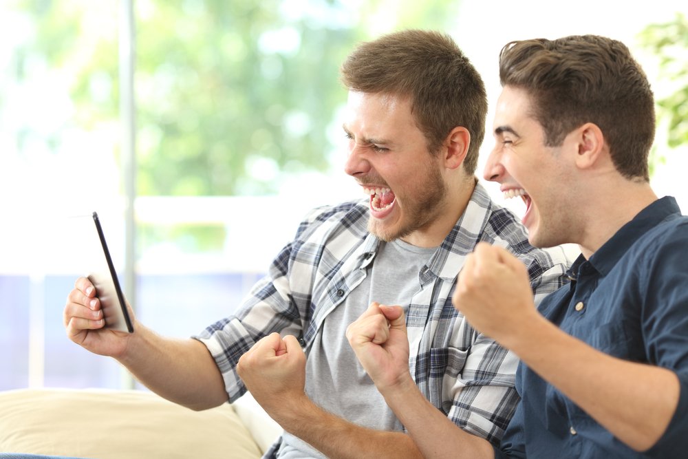 Dos emocionados amigos o compañeros de habitación viendo la televisión en línea en una tableta sentados en un sofá en la sala de estar en casa. I Foto: Shutterstock