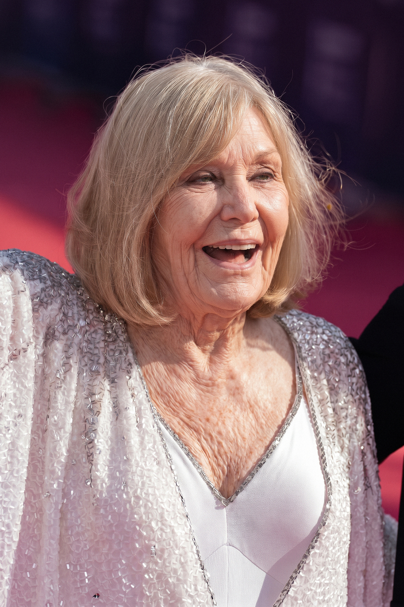 Kim Novak sonríe ampliamente con una chaqueta plateada brillante sobre un vestido blanco con cuello en V, y su corte de pelo rubio a lo garçon le enmarca el rostro sobre un fondo rosa y morado del evento. | Fuente: Gemini
