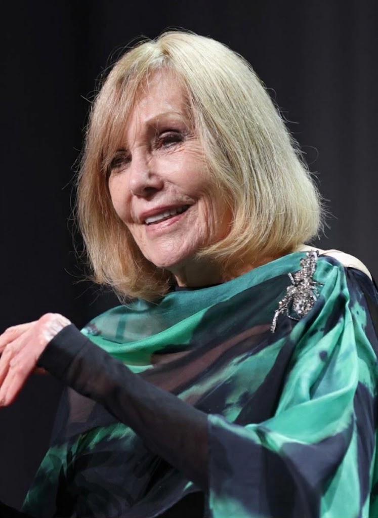 Kim Novak sonríe durante una aparición en el escenario con un vestido holgado verde y negro, con mangas transparentes y un broche brillante que le da un toque especial a su look. | Fuente: Gemini