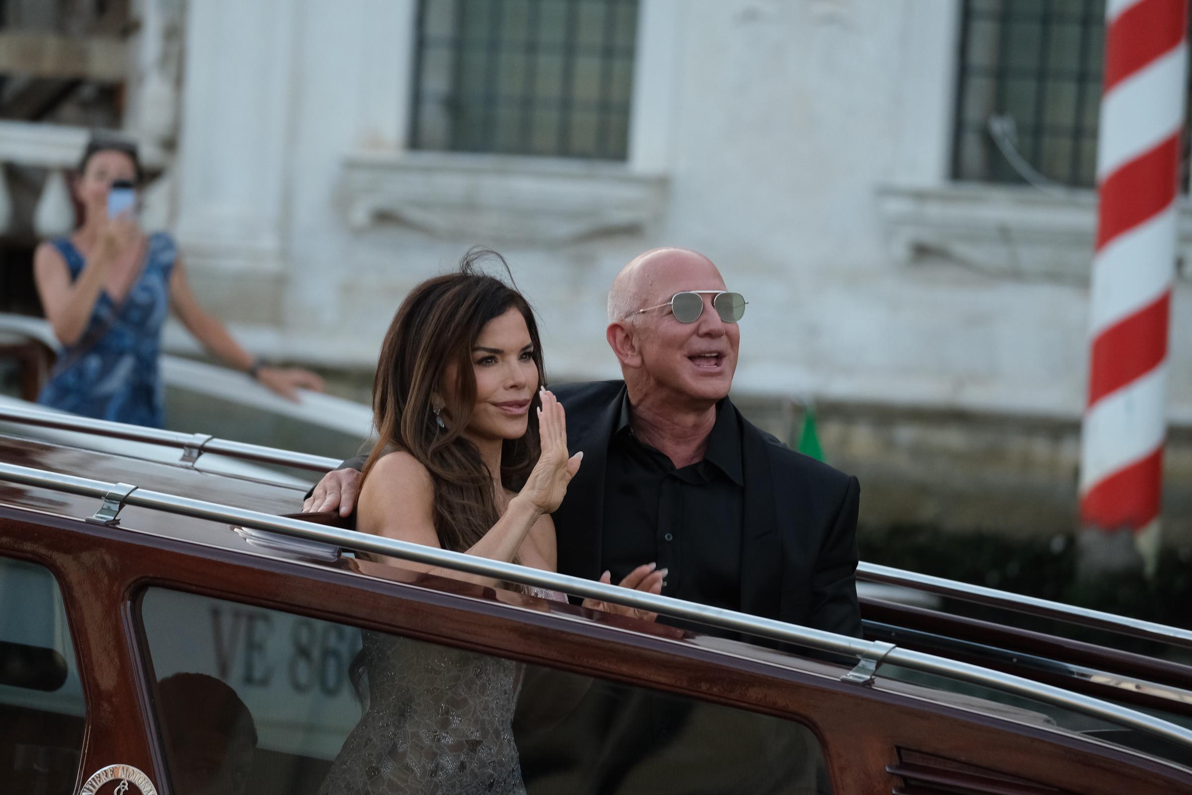 Lauren Sánchez levanta la mano en un suave gesto de saludo mientras Jeff Bezos sonríe a su lado, ambos de pie en el barco, mientras una mujer al fondo capta el momento con su teléfono cerca de un poste a rayas rojas y blancas.