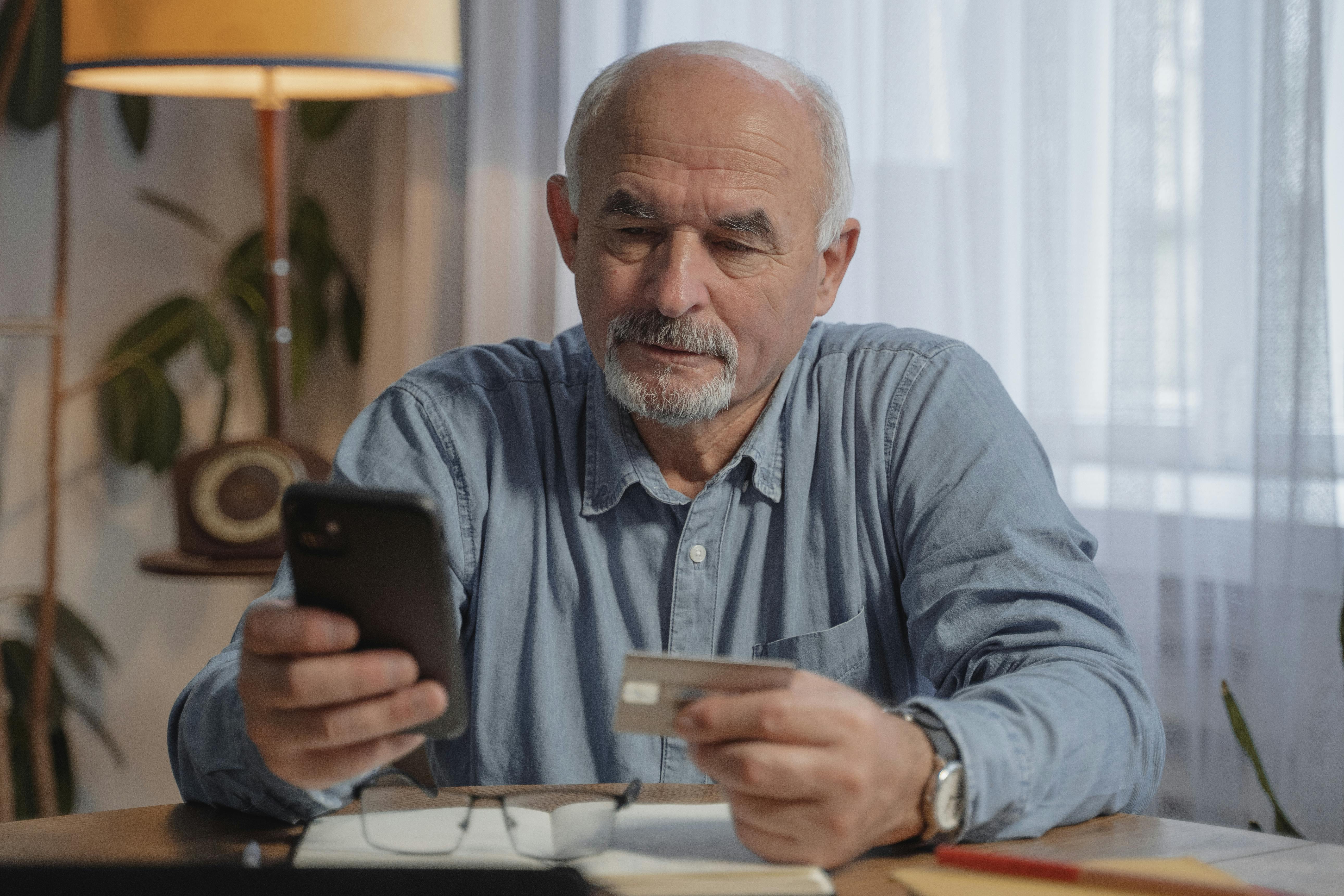 Un hombre mayor con el teléfono en una mano y una tarjeta bancaria en la otra | Foto: Pexels