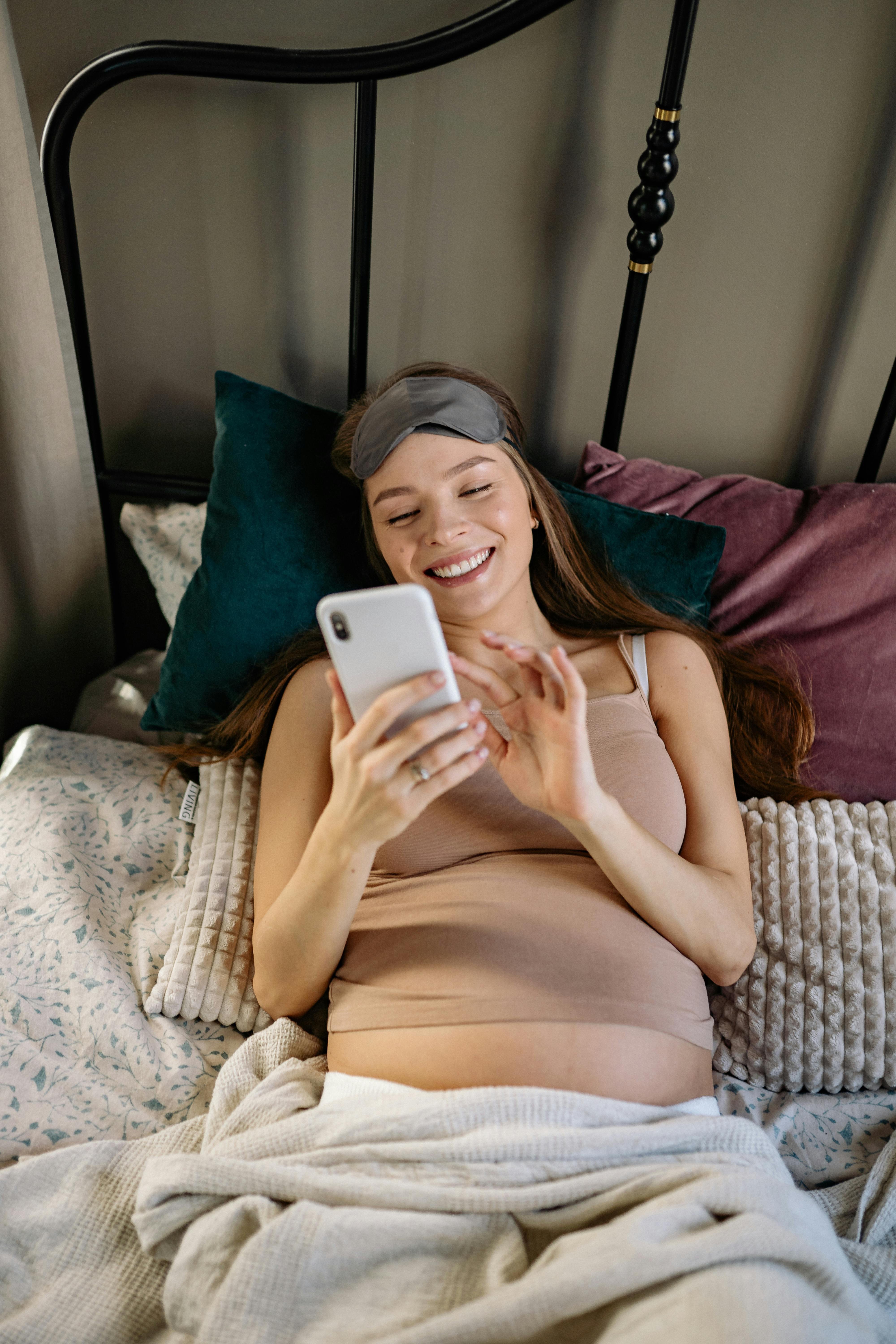 Una embarazada sonriente leyendo el móvil en la cama | Foto: Pexels