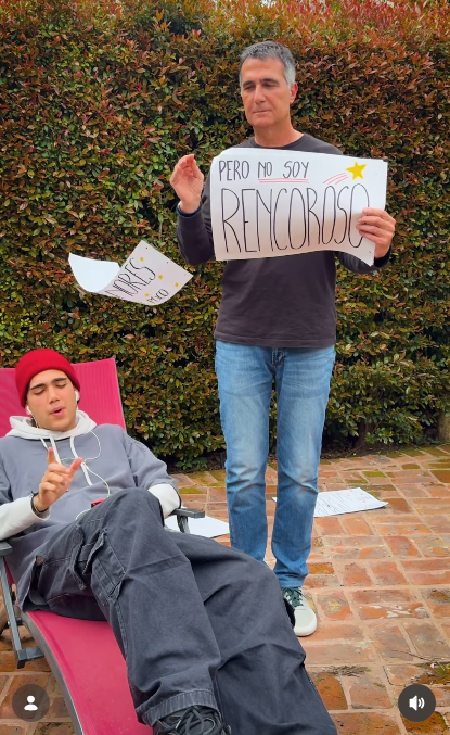 El periodista Antonio Laje exhibió la cartulina con la inscripción "PERO NO SOY RENCOROSO" mientras dejaba caer al suelo el cartel anterior. En primer plano, su hijo Ezequiel (Leish) gesticulaba con la mano al ritmo de su canción, vestido con un buzo gris holgado, pantalones cargo negros y un gorro rojo. | Fuente: Instagram/leish.ar