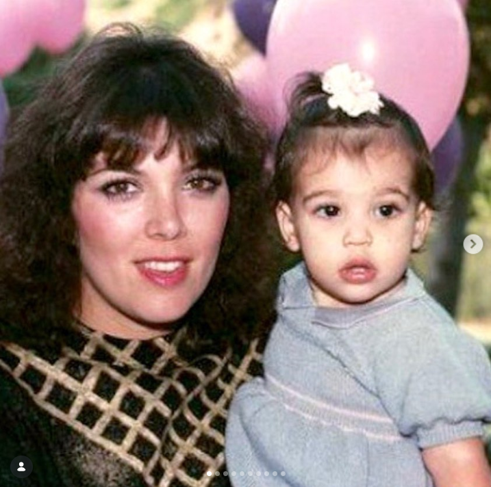 Una joven Kris Jenner posa con su hija Kourtney Kardashian cuando era un bebé, abrazándola con cariño mientras posan delante de un fondo de globos rosas. Kris lleva una camiseta estampada, mientras que Kourtney lleva un conjunto gris claro y lleva una florecita blanca en el pelo. | Fuente: Instagram/krisjenner