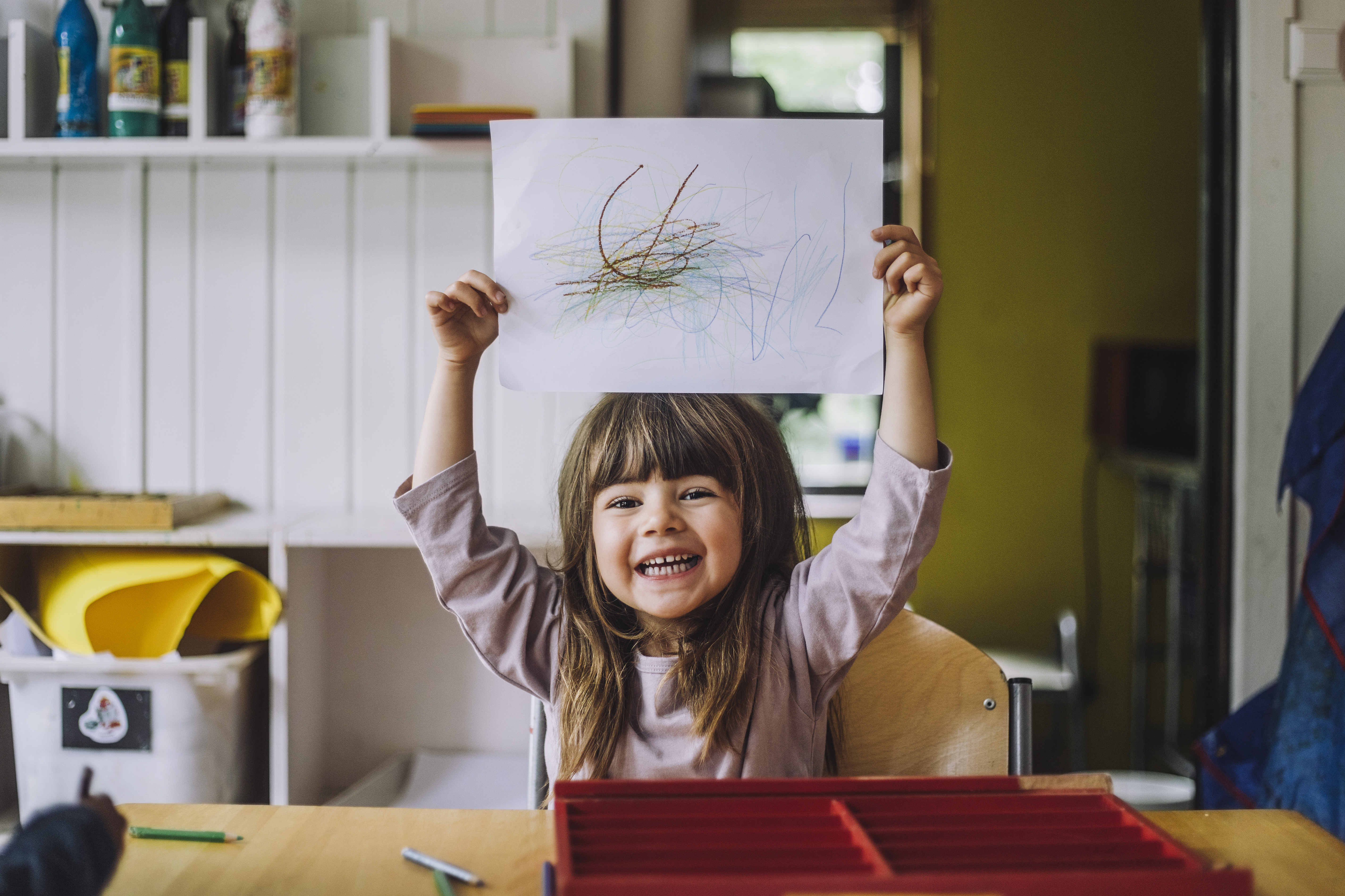 Niña feliz mostrando su garabato de lápiz de color sobre papel en la guardería | Foto: Getty Images