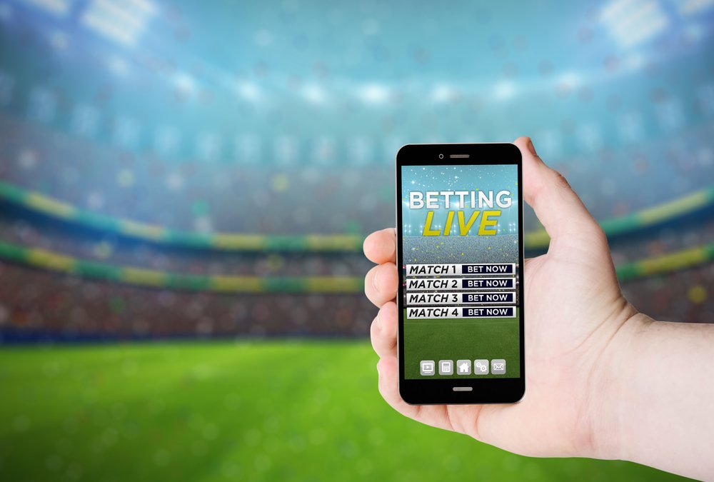 Mano sostiene un teléfono con apuestas en línea en una pantalla en el fondo de un estadio. I Foto: Shutterstock