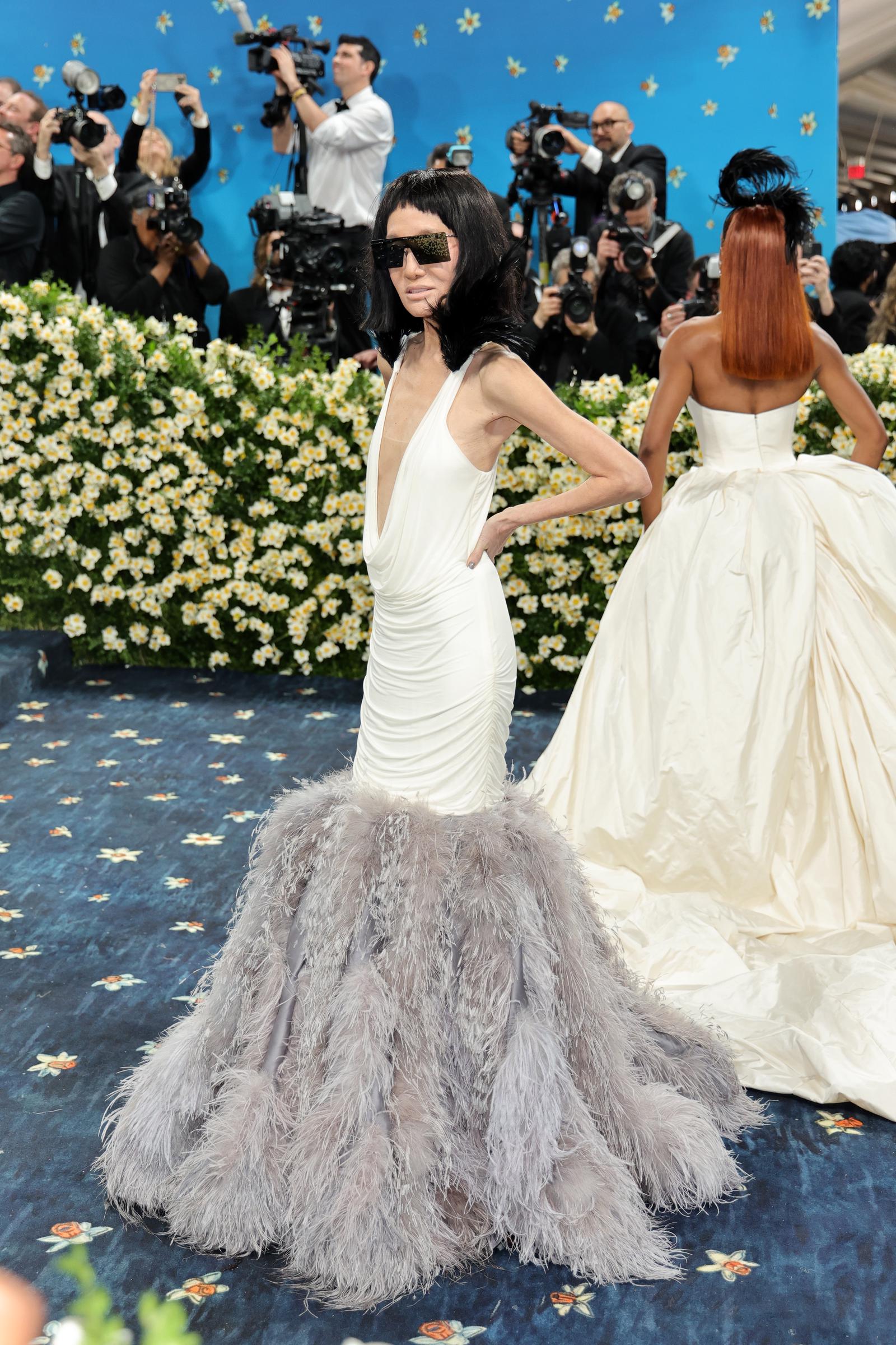 Con las cámaras parpadeando detrás de ella, Vera Wang posa de perfil, mostrando el drapeado esculpido del vestido y la voluminosa falda de plumas sobre un fondo floral.