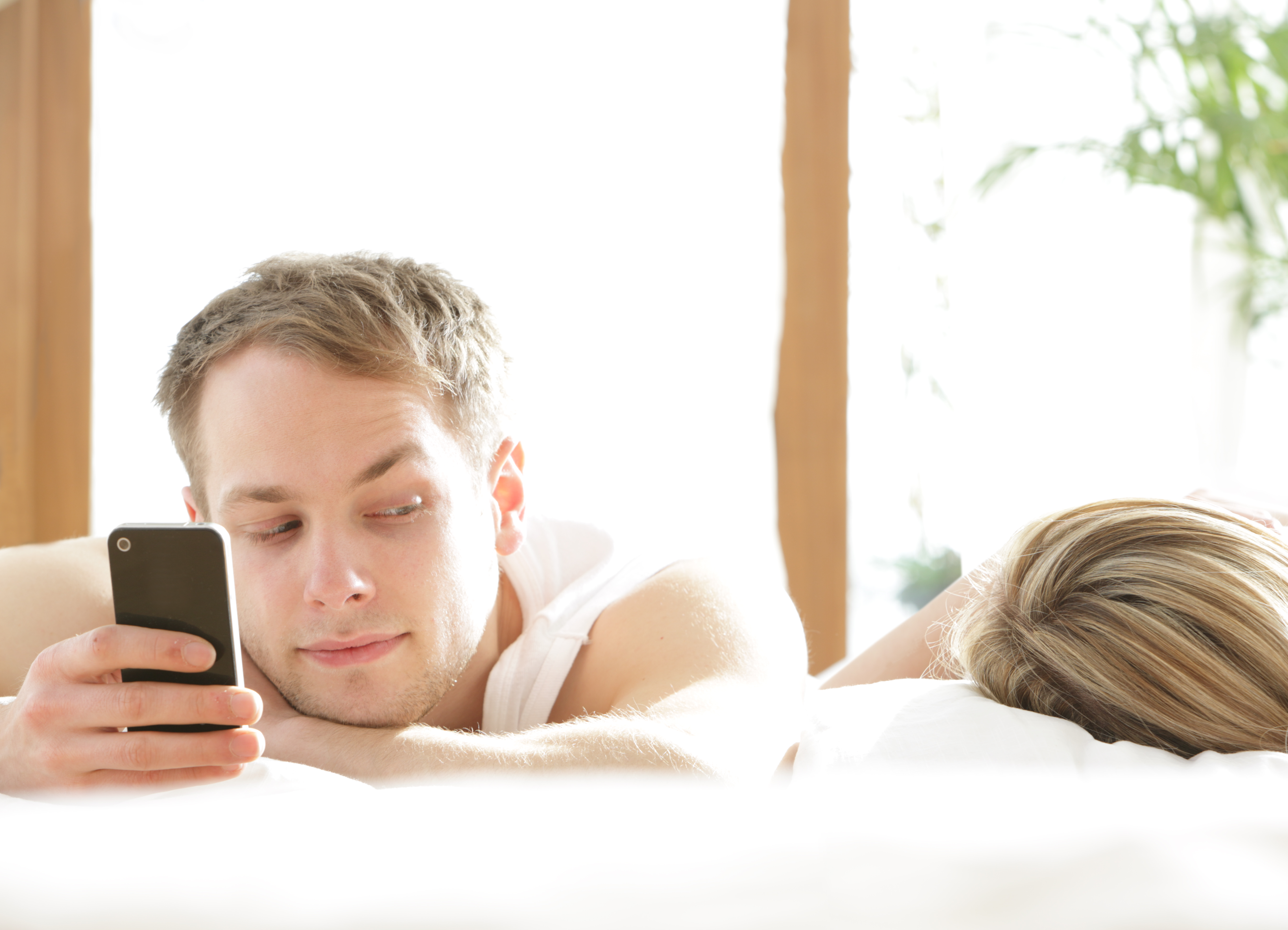 Hombre con teléfono en la cama, mirando a mujer dormida | Fuente: Getty Images