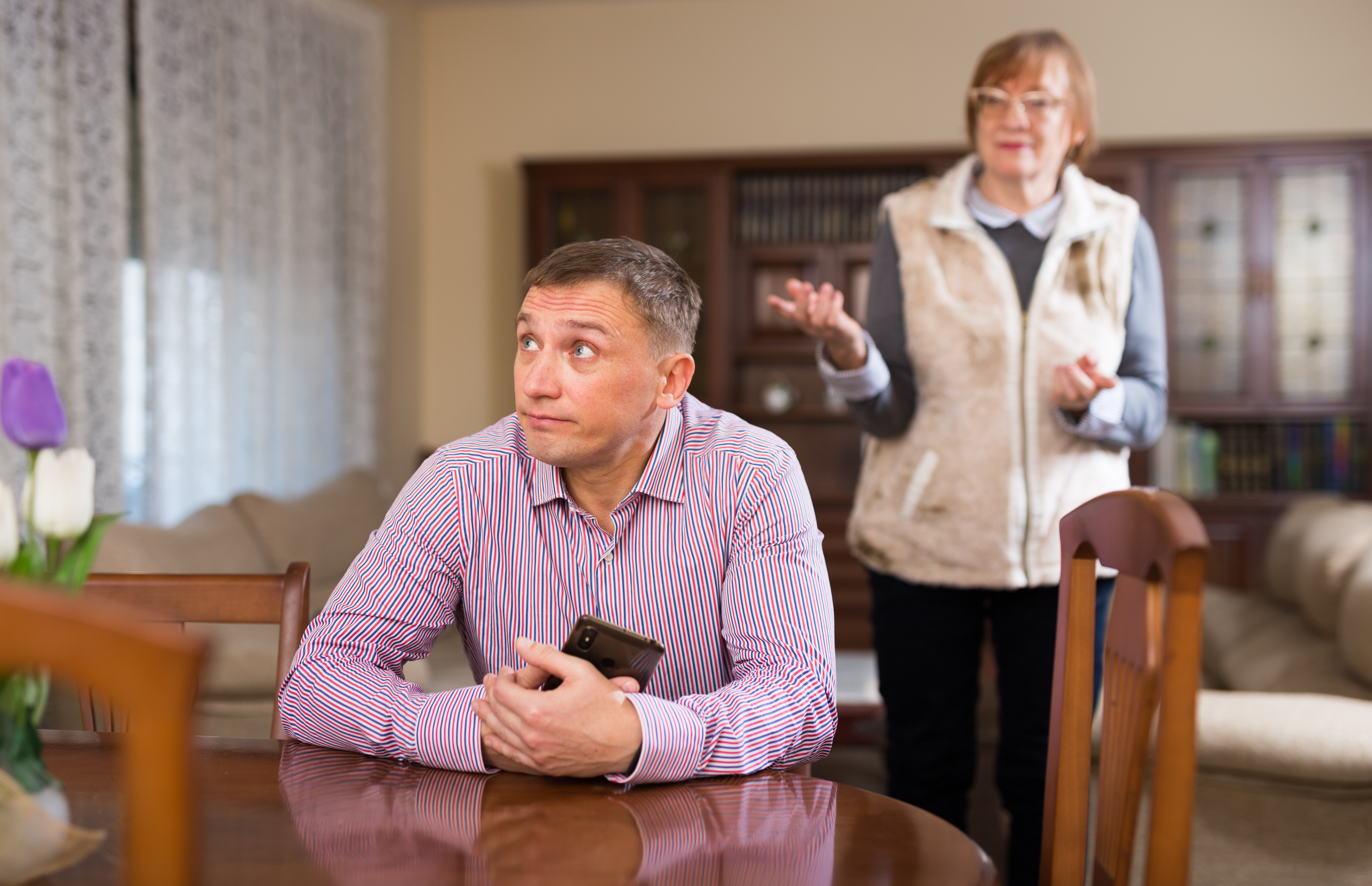 Un hombre joven agarra su teléfono y mira hacia otro lado sentado a una mesa mientras una señora mayor intenta hablarle en segundo plano. | Foto: Shutterstock