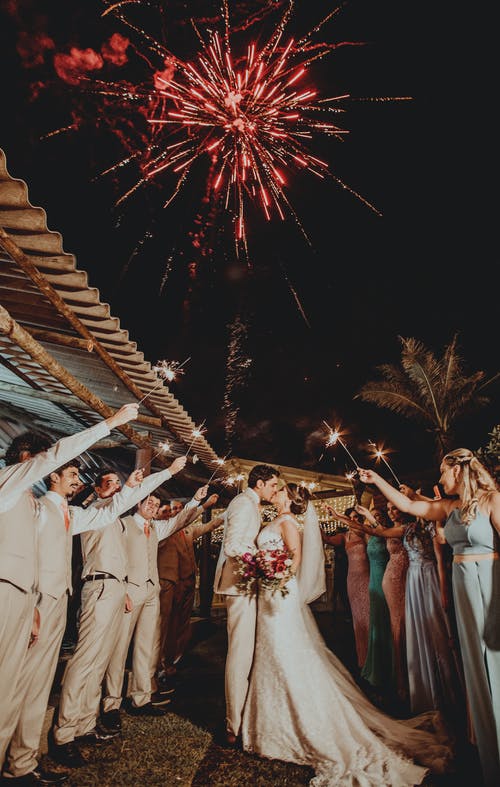 Una pareja de recién casados rodeados de amigos con fuegos artificiales en el fondo. | Foto: Pexels