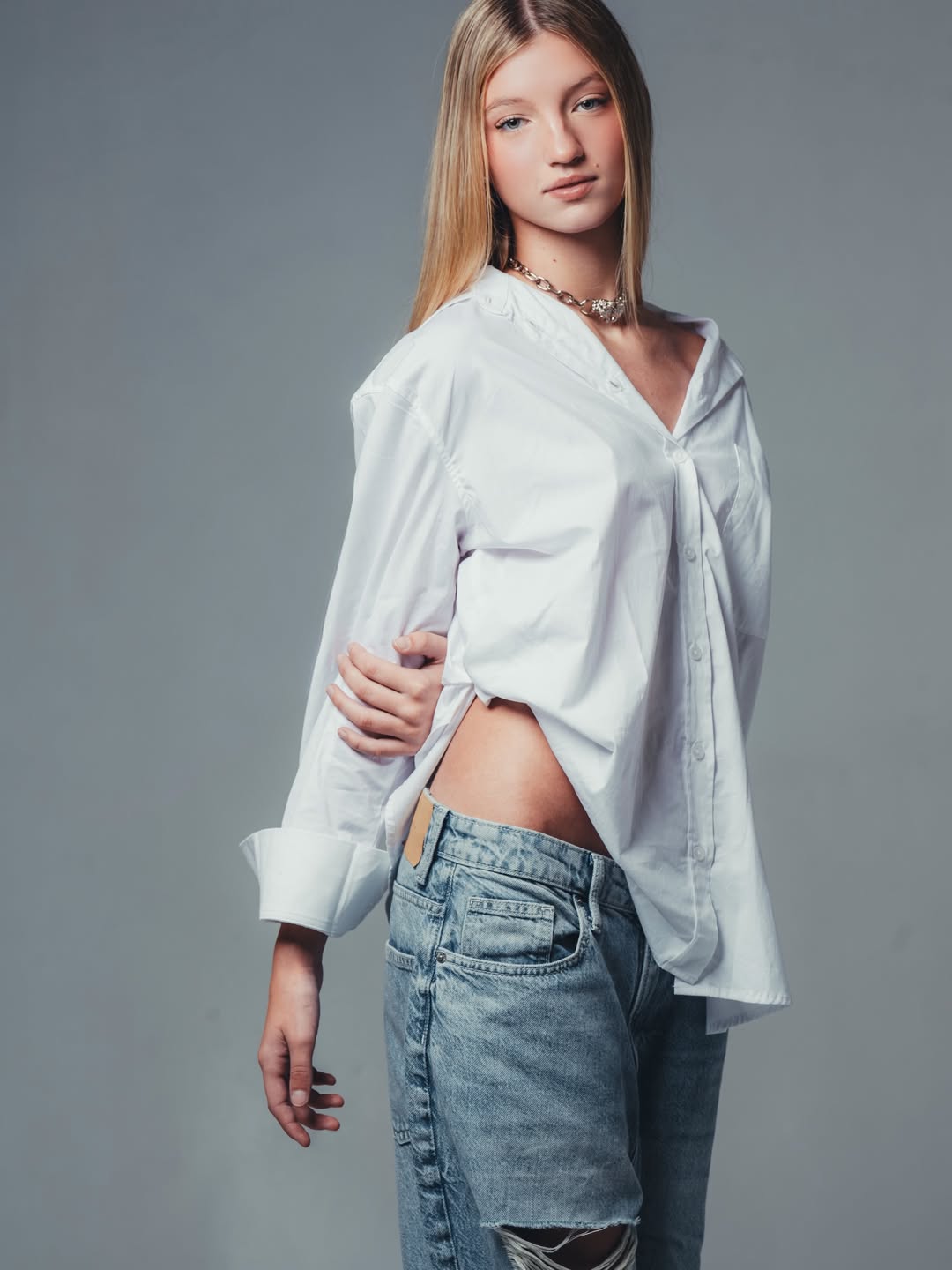 Allegra Cubero posa con camisa blanca holgada y jeans, con collar visible y mirada suave frente a fondo gris.