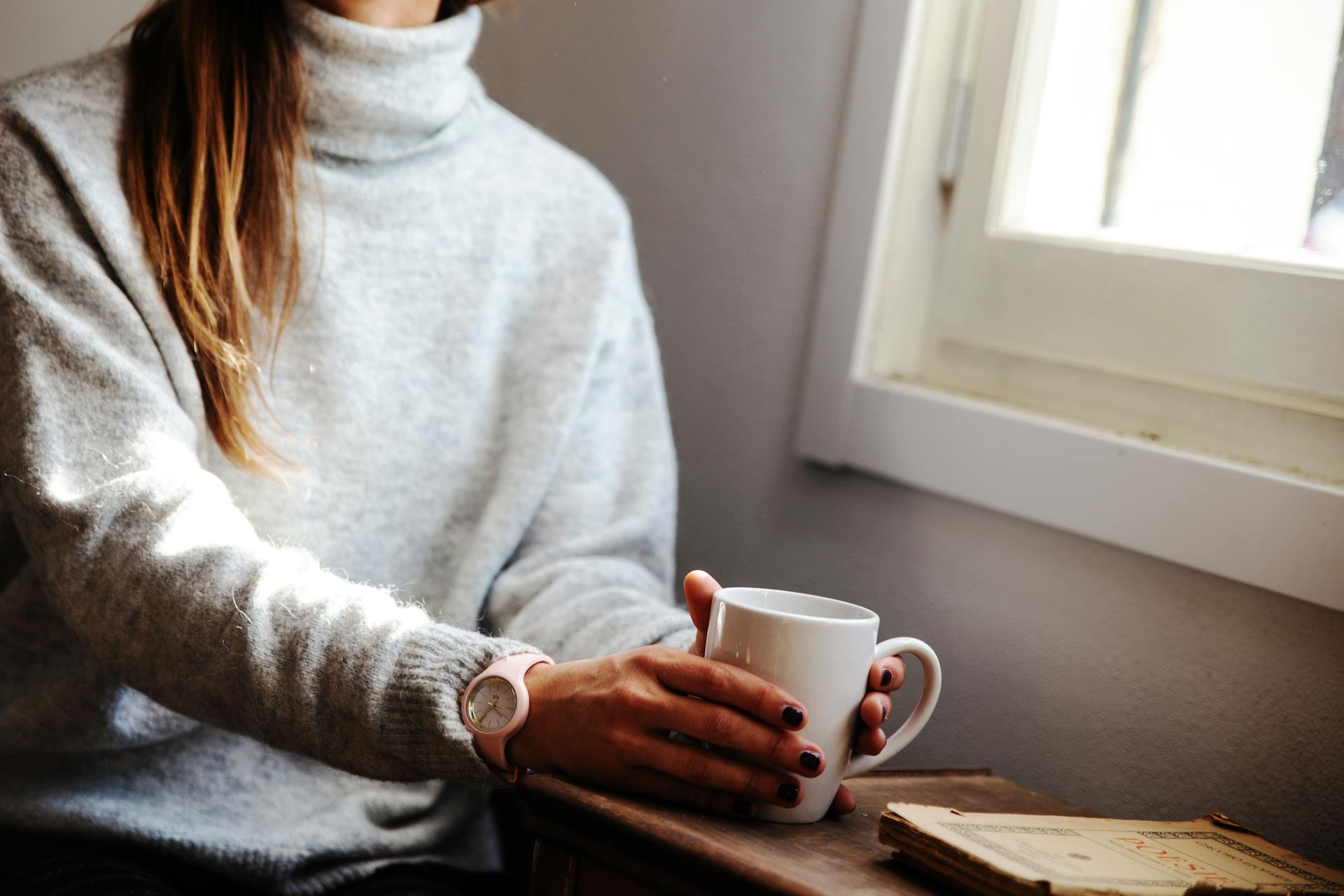Una mujer con una taza de café en la mano y sentada junto a la ventana | Fuente: Pexels