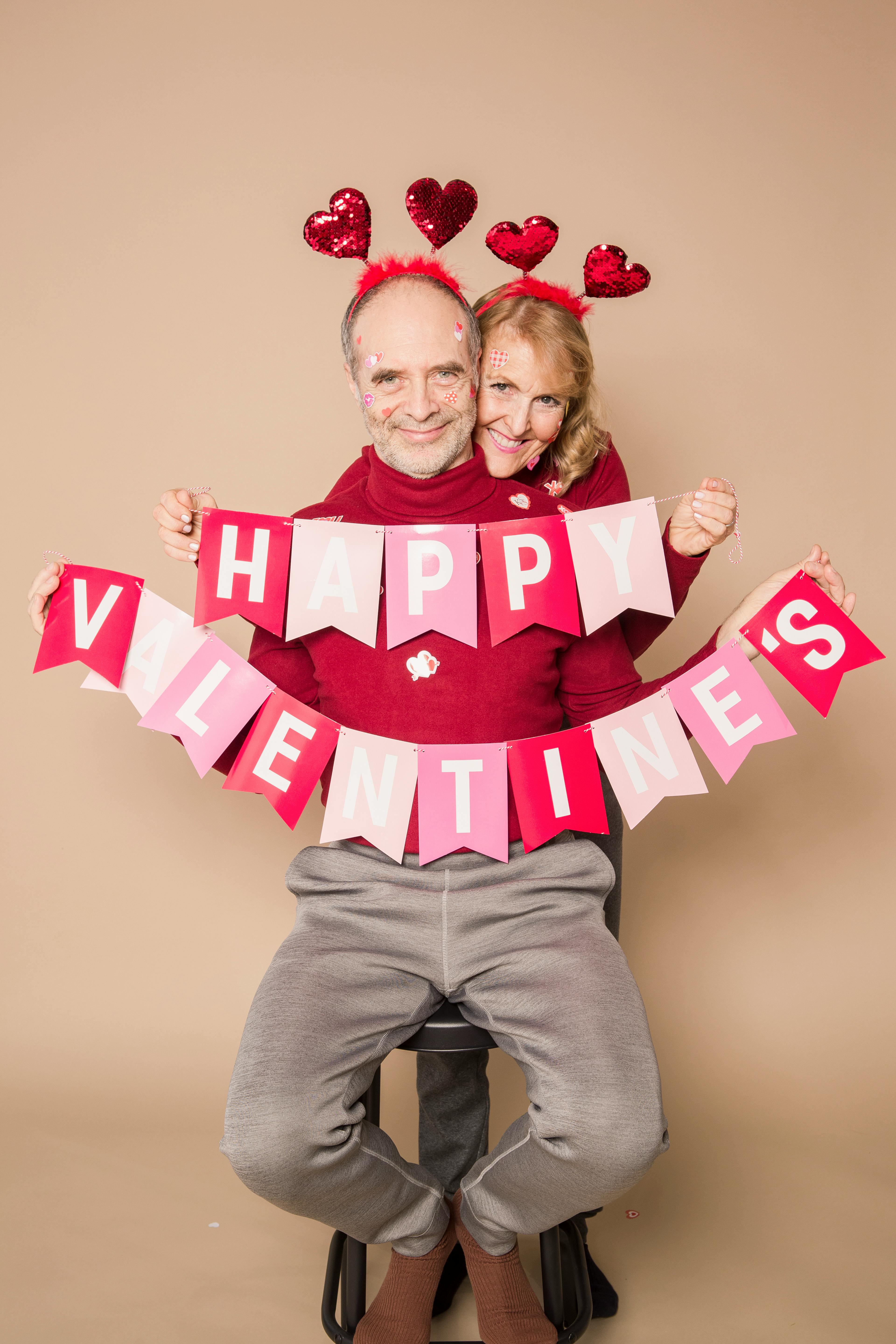 Una pareja con trajes a juego sujetando una decoración colgante que dice "Feliz San Valentín". | Fuente: Pexels