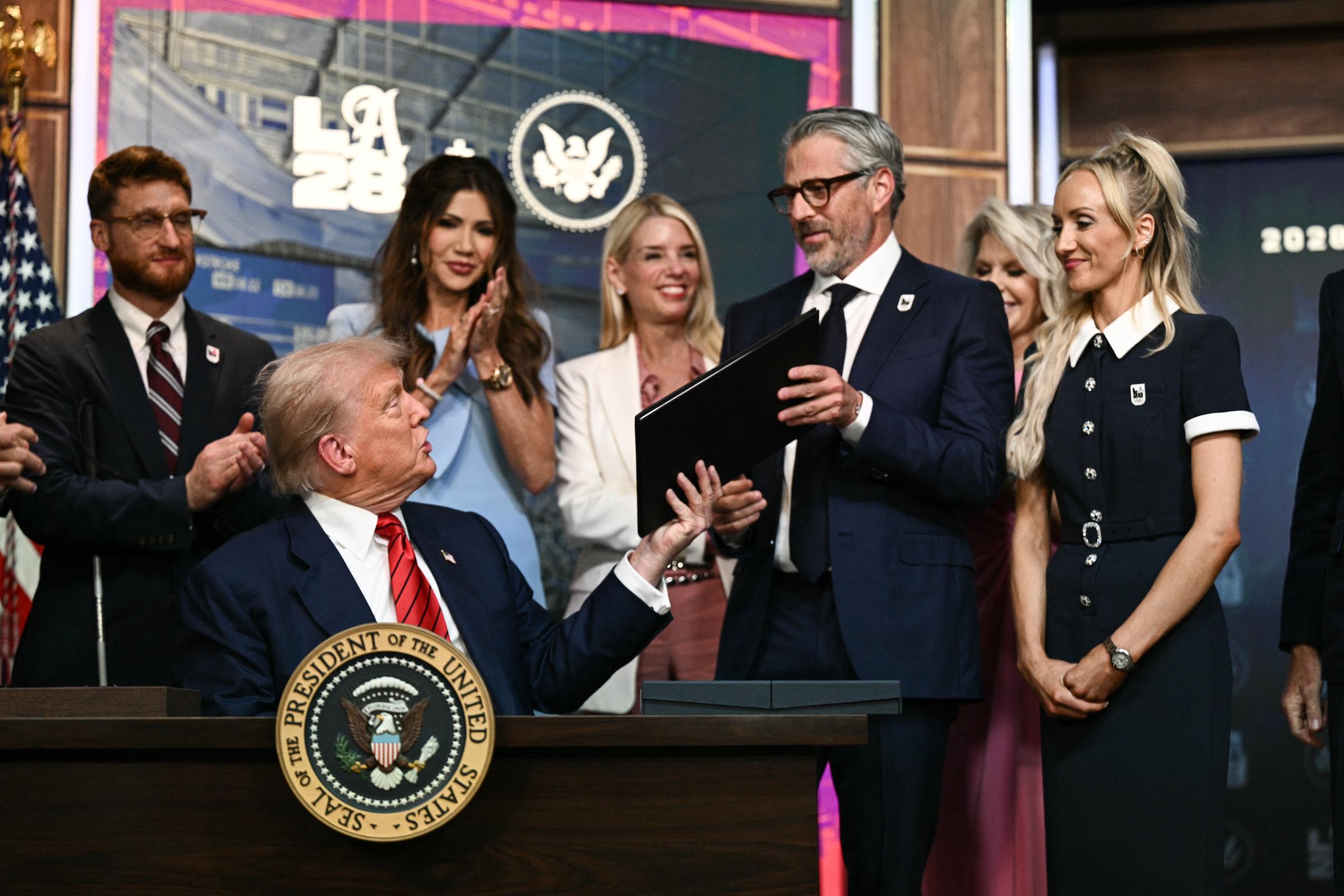 Donald Trump parece estar hablando mientras le entrega al presidente del comité organizador de Los Ángeles 2028, Casey Wasserman, una copia de la orden ejecutiva firmada, mientras los asistentes observan con sonrisas y aplausos.