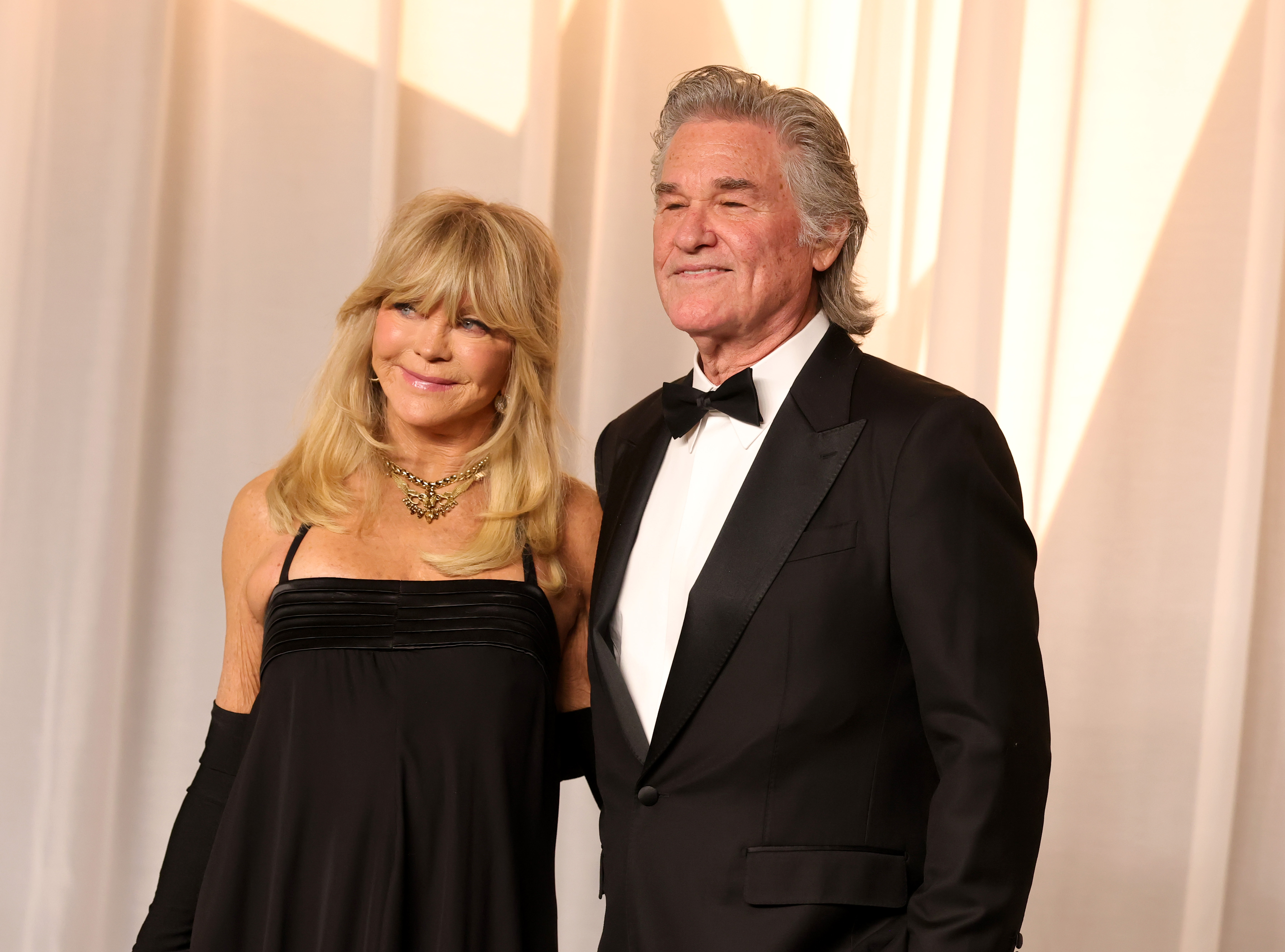Hawn y Russell posan juntos con elegantes atuendos de gala: el vestido de tirantes de Hawn, los guantes largos, las joyas llamativas y su voluminosa melena rubia contrastan con el esmoquin a medida de Russell, su impecable camisa blanca y su pelo gris peinado hacia atrás.