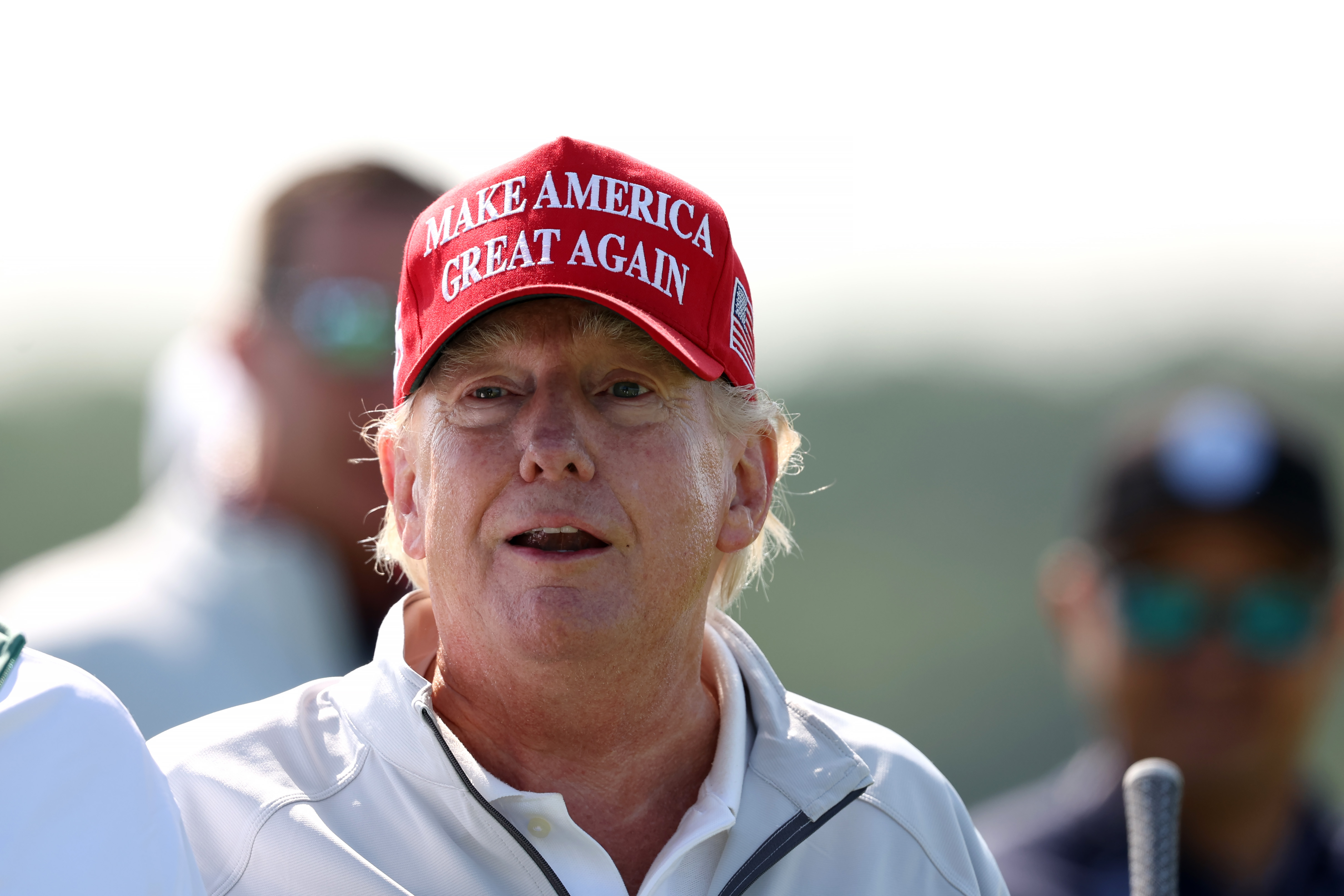 En el LIV Golf Invitational en Sterling, Virginia, el 25 de mayo, Donald Trump está de pie bajo el sol con una gorra roja de "Make America Great Again" y una chaqueta blanca con cuello. Sus mejillas y nariz se ven intensamente enrojecidas, mientras que su cuello luce mucho más claro.