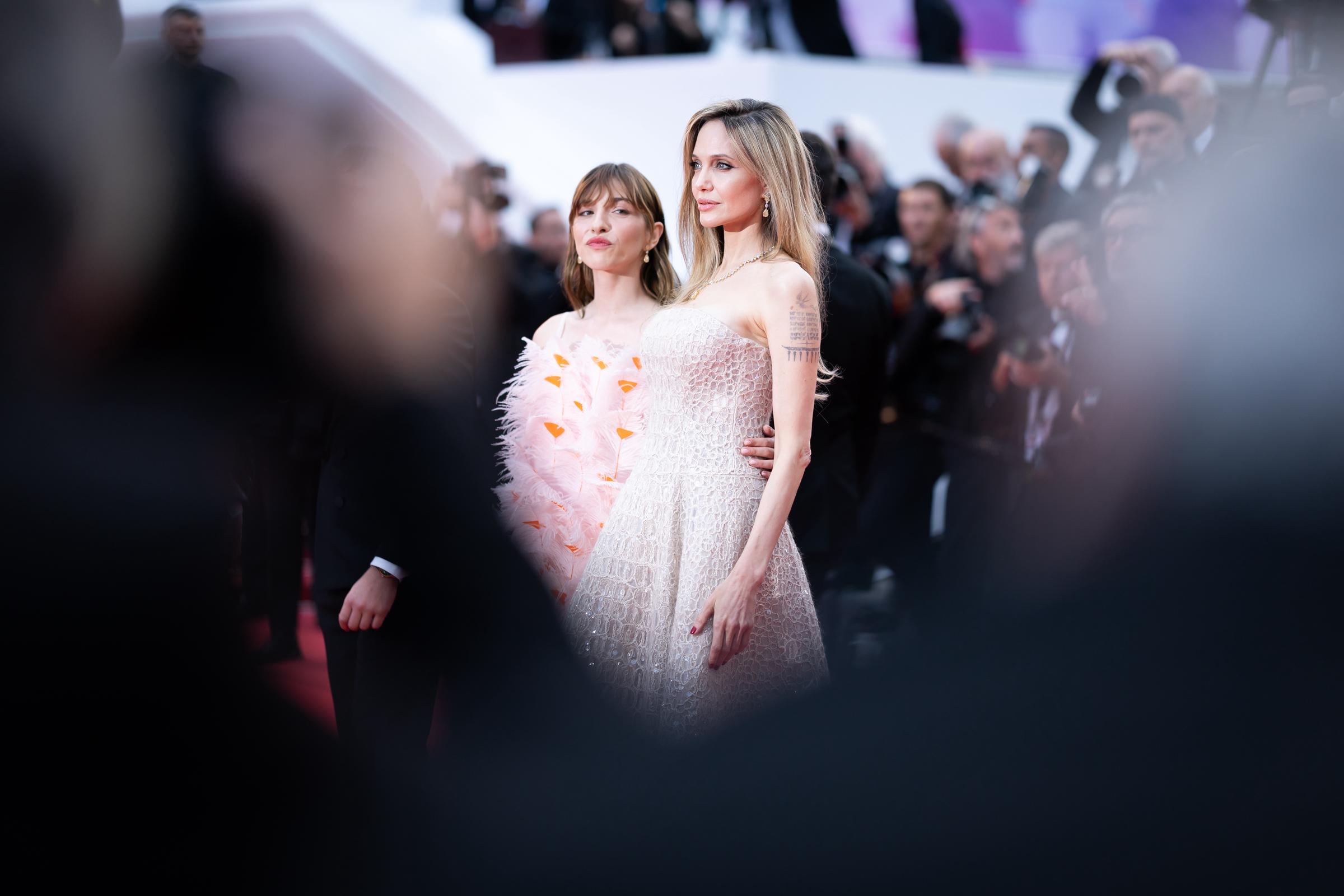 Enmarcadas por siluetas difuminadas en primer plano, Angelina Jolie y Marie Colomb comparten un momento impactante en la alfombra roja. Jolie se mantiene erguida con su vestido de lentejuelas de Cucinelli, mientras que Colomb deslumbra con un minivestido de plumas de inspiración floral, ambas mujeres acaparando la atención desde todos los ángulos.