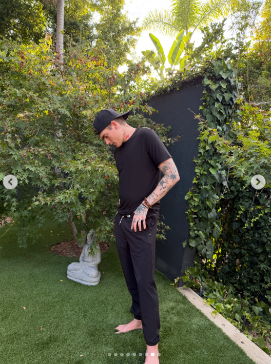 Presley Gerber mira hacia abajo mientras se ajusta los pantalones negros, de pie descalzo en el jardín con la gorra puesta al revés y los tatuajes a la vista entre la exuberante vegetación. | Fuente: Instagram/presleygerber